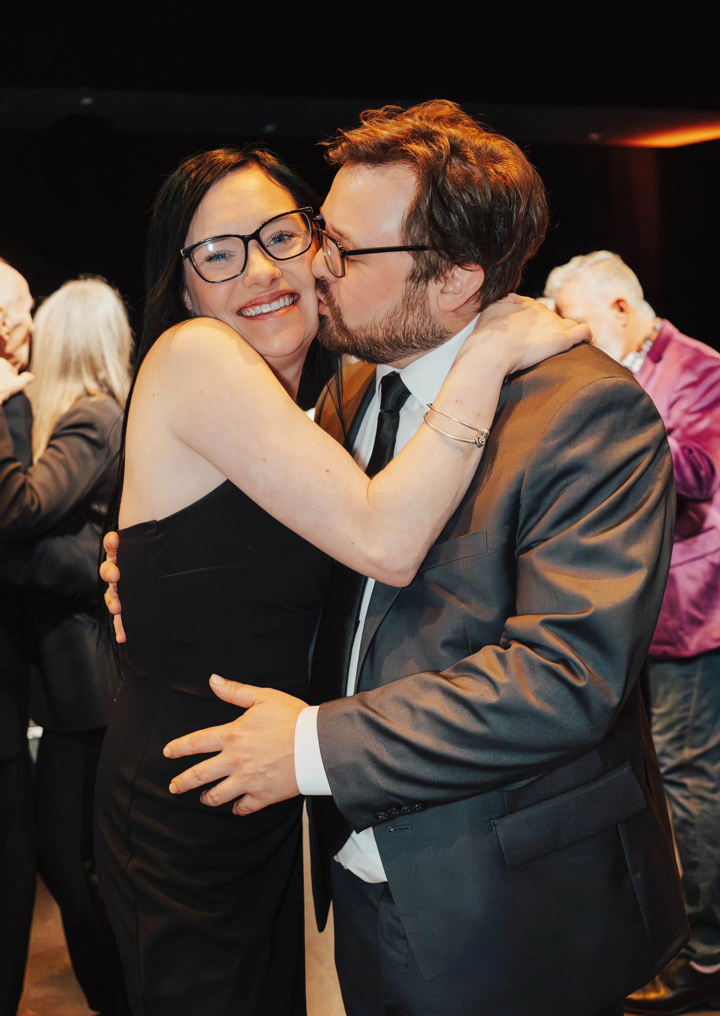 A woman and a man hugging at a formal event. The woman is smiling, wearing glasses and a black dress. The man is kissing her cheek, wearing glasses and a black suit with a tie. Other people are visible in the background.