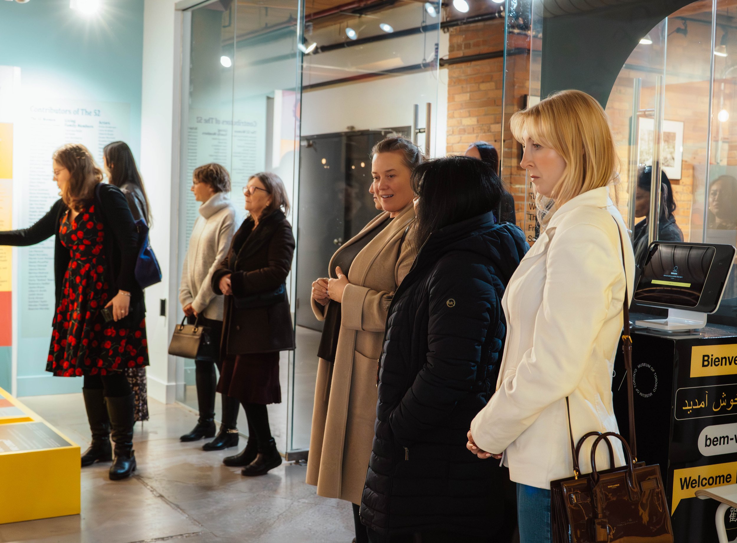 A group of women are standing in a line inside a museum or exhibit space, observing displays on the wall and a computer station visible on the right. The setting has glass partitions and brick walls, with some visitors looking attentively at the exhi