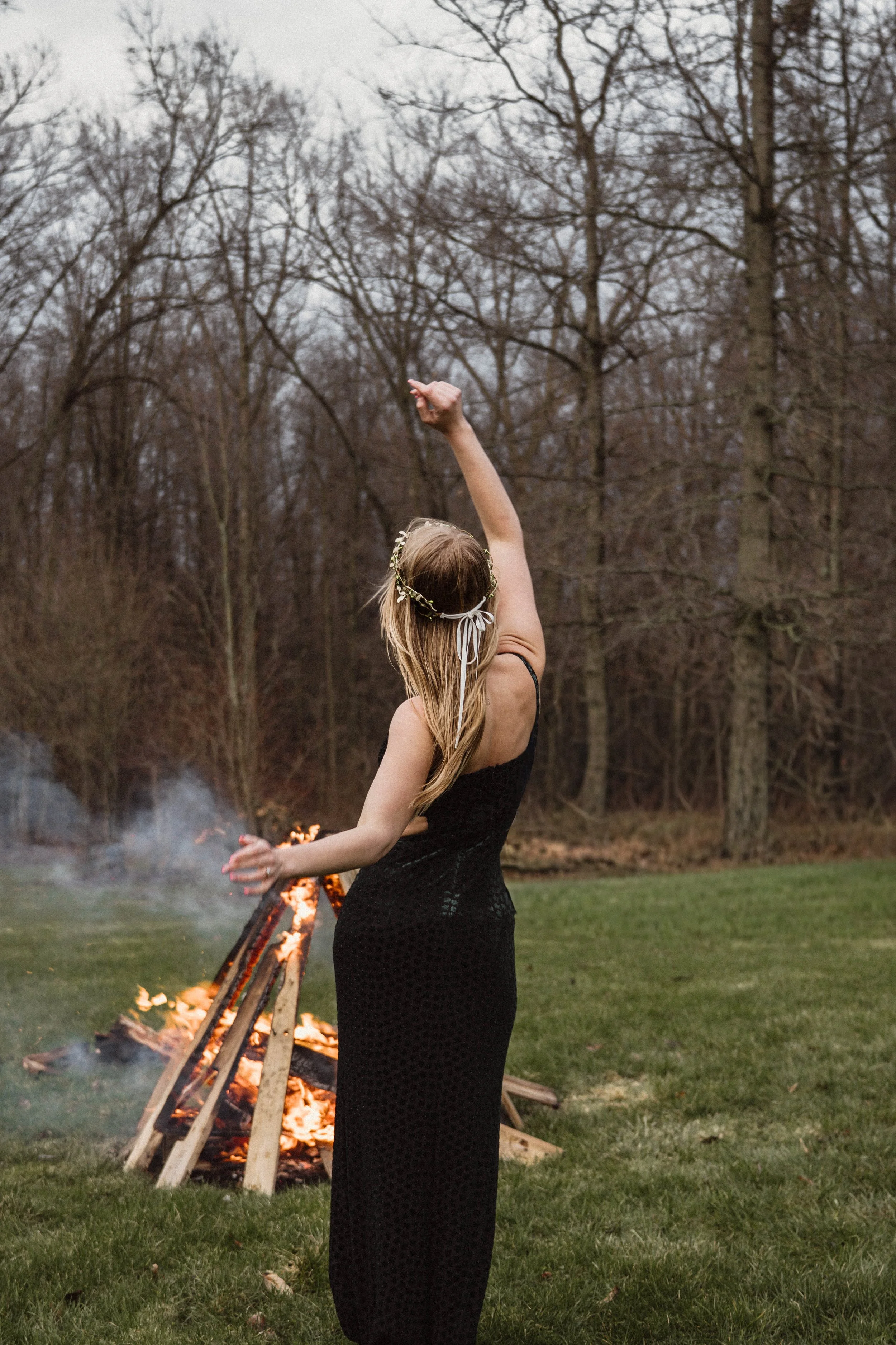 A woman in a black dress with a floral headband performing a dance outdoors near a fire pit in a wooded area.