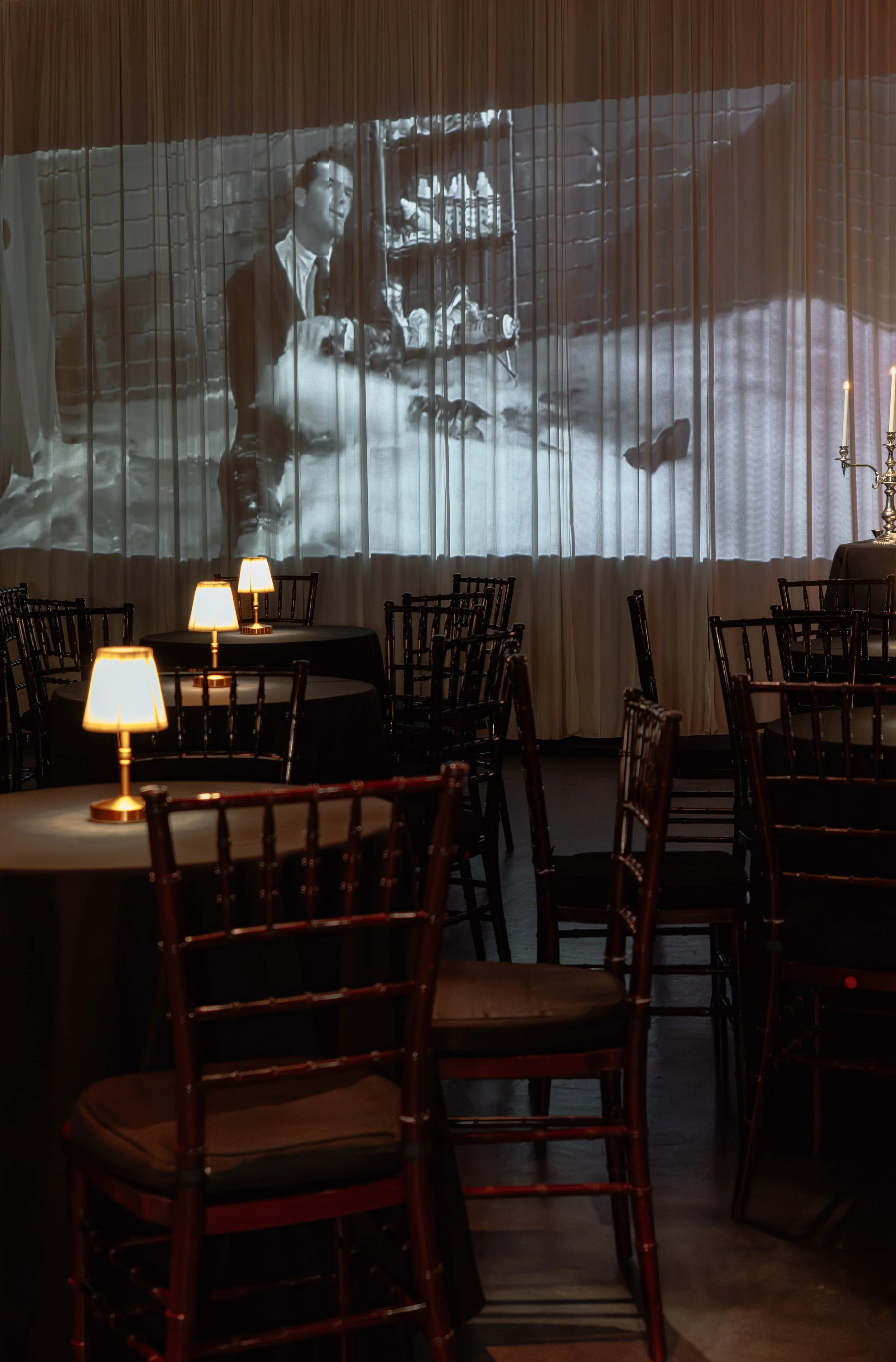 Indoor event space with round tables, black tablecloths, and wooden chairs with cushions, illuminated by small table lamps. A large screen or curtain at the back of the room shows a black-and-white film or photograph of a man in formal attire.