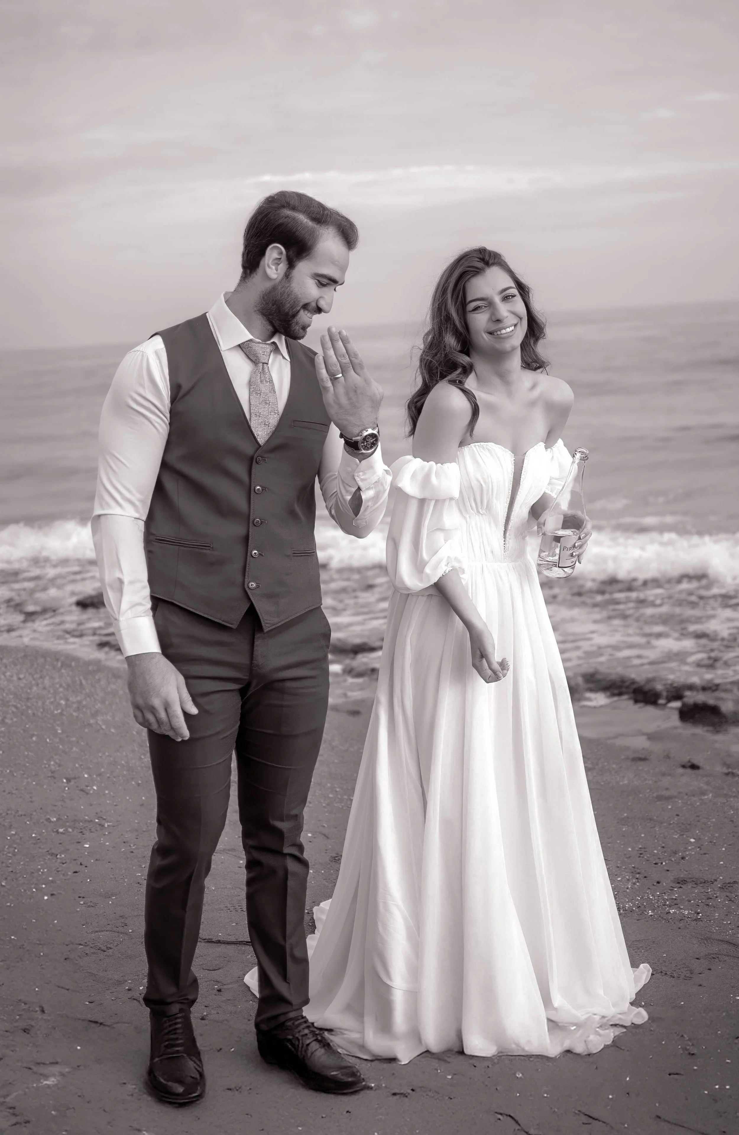 A black and white photo of a couple on the beach, with the man wearing a vest and tie, and the woman in a flowing white dress holding a drink, smiling at the camera.