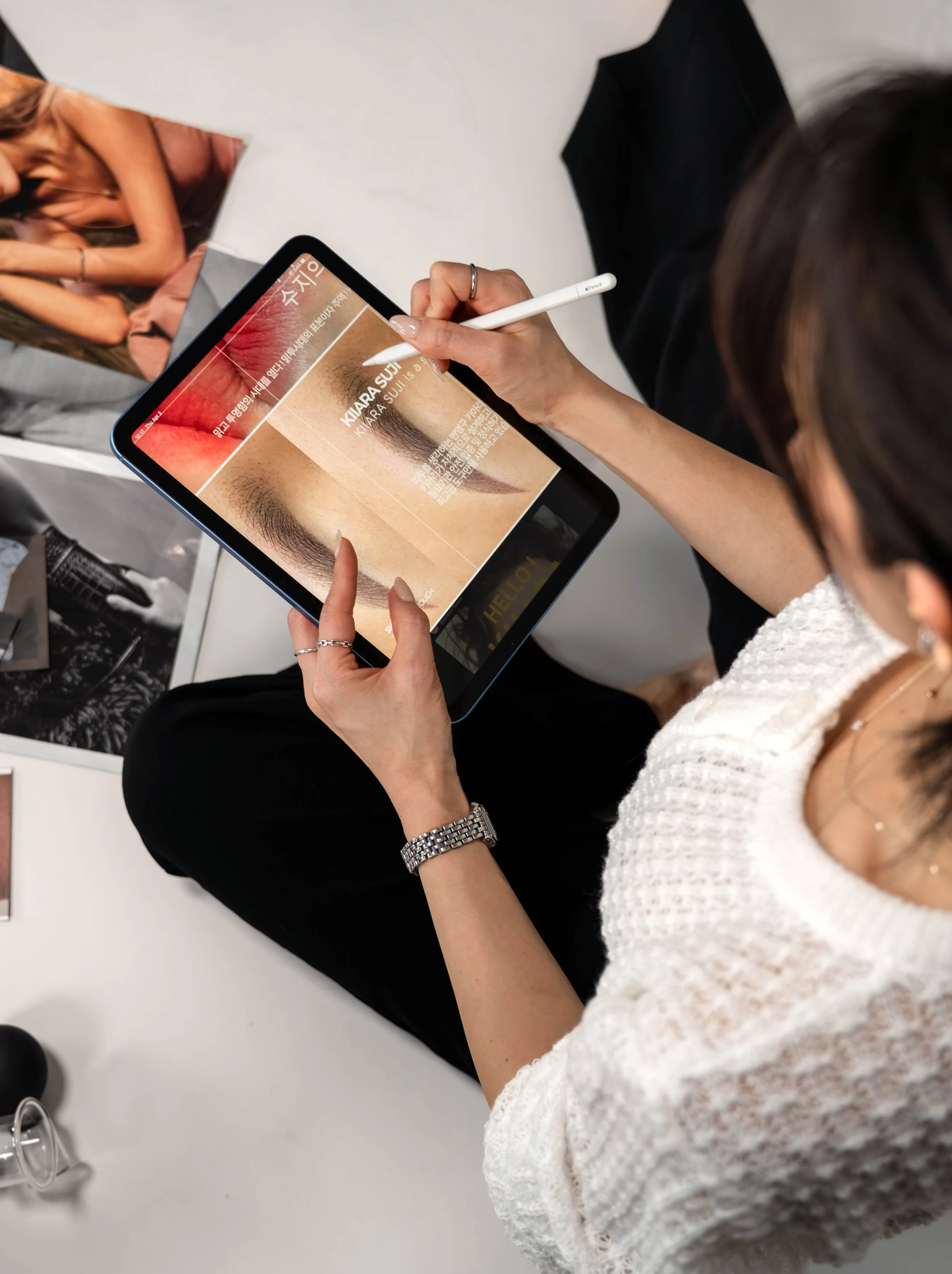 A woman sitting on a white surface is using a stylus to navigate a digital tablet displaying information about eyebrows, possibly related to cosmetic procedures or beauty services. She is wearing a white textured top, a silver watch, and rings, with 