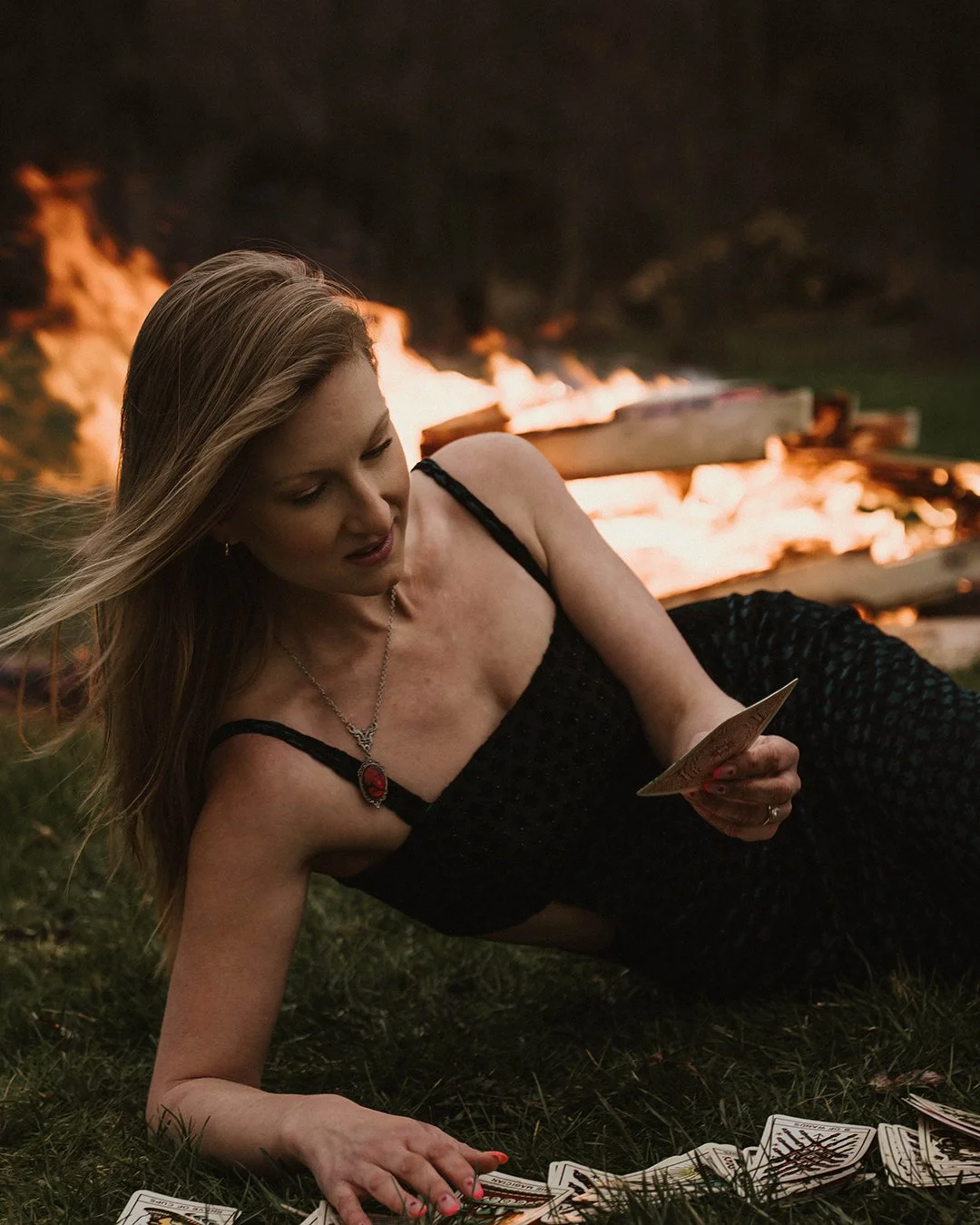 A woman with long blonde hair lying on the grass, reading tarot cards, with a large fire burning in the background.