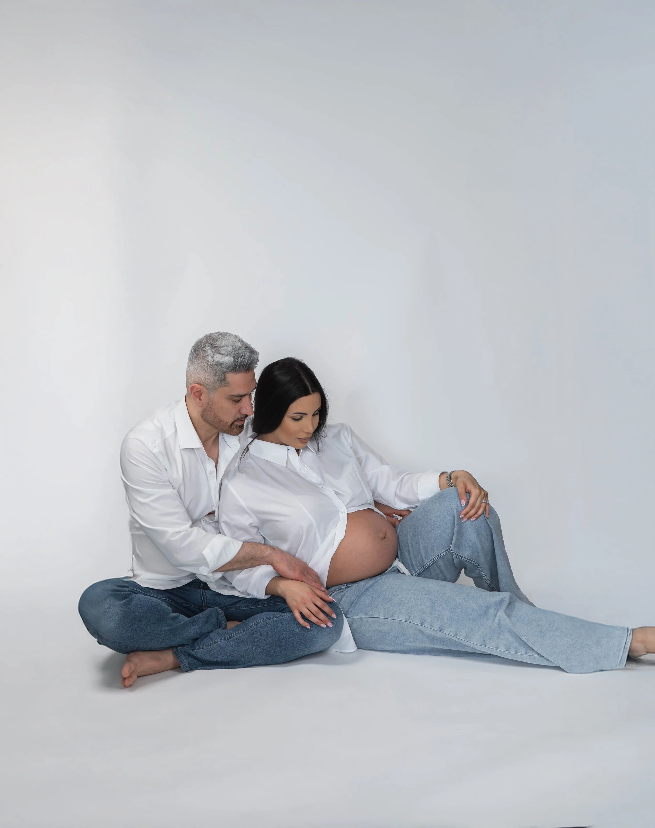 A man and woman sitting on the floor with a pregnant woman, the woman is holding her belly and the man is touching her knee, all wearing white shirts and jeans, in front of a plain white wall.