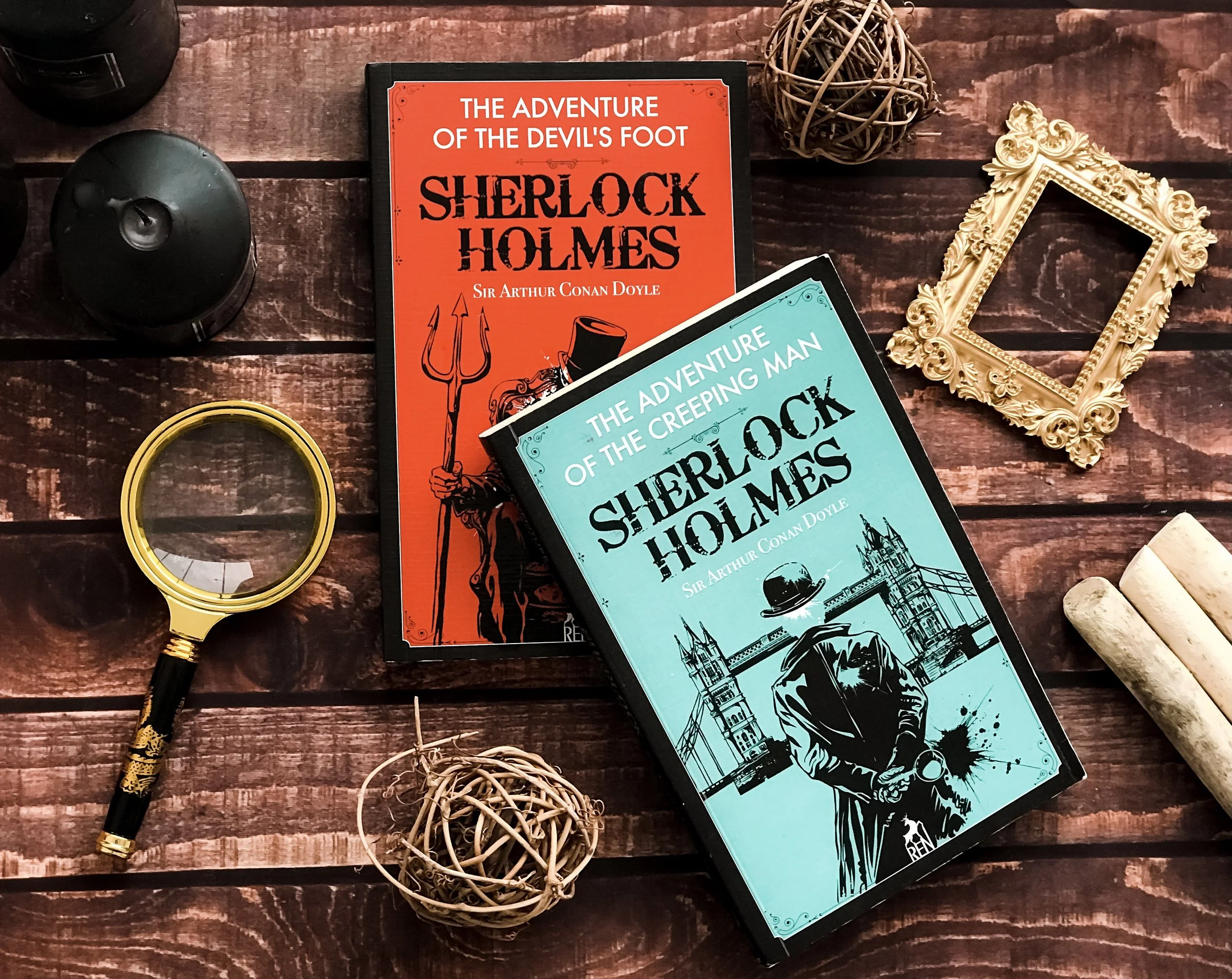 Two books titled "The Adventure of the Devild's Foot" and "The Adventure of the Creeping Man" by Sherlock Holmes, on a wooden surface with decorative items including a magnifying glass, a small woven ball, a gold ornate picture frame, black and white