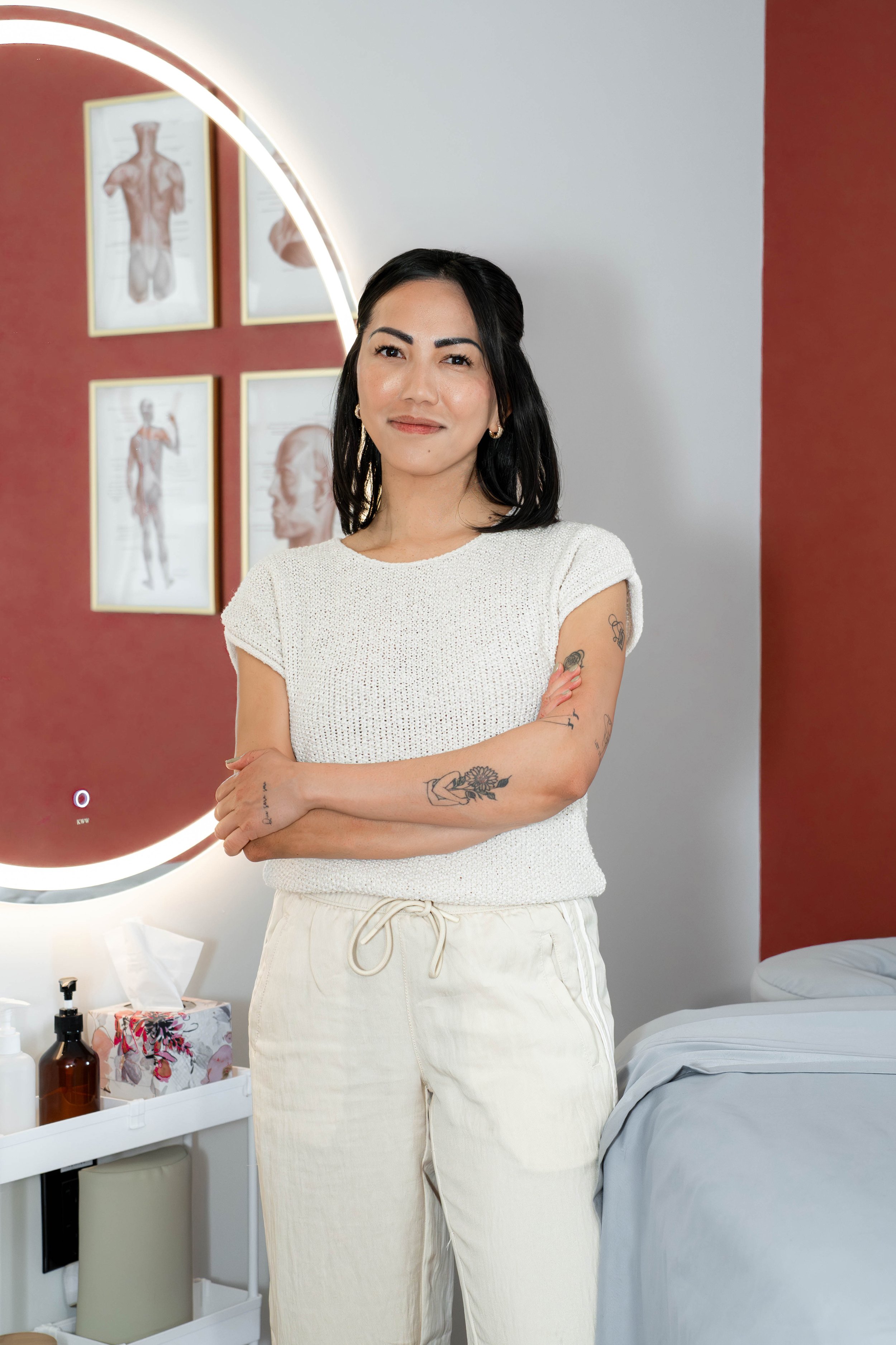A woman with black hair, tattoos on her arms, wearing a white knitted top and cream-colored pants, standing inside a room with a red wall and framed anatomical sketches in the background.