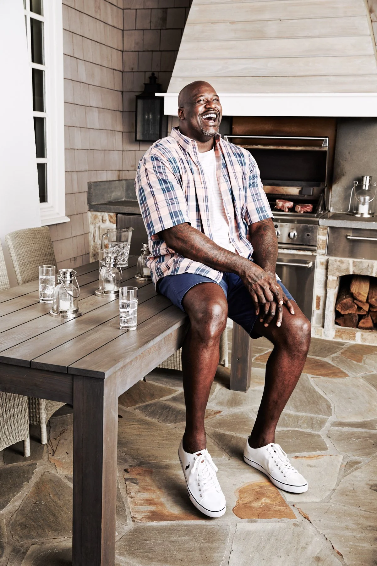 Person sitting on a table outdoors with grill and patio area, wearing a plaid shirt, shorts, and sneakers.