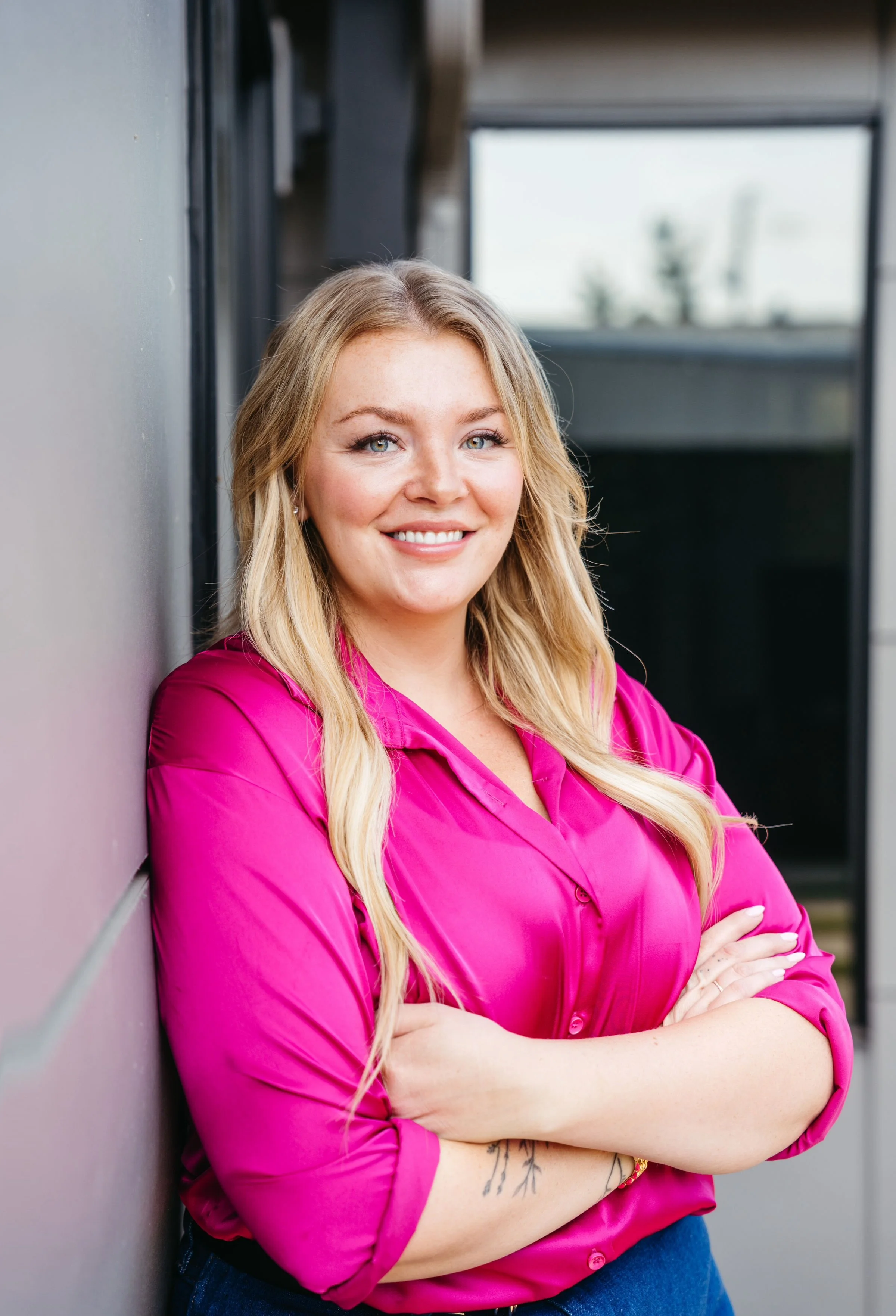 A woman in a pink blouse sitting at a desk in an office, smiling at the camera. There is a printer and a stack of papers behind her, with a large round clock on the wall.