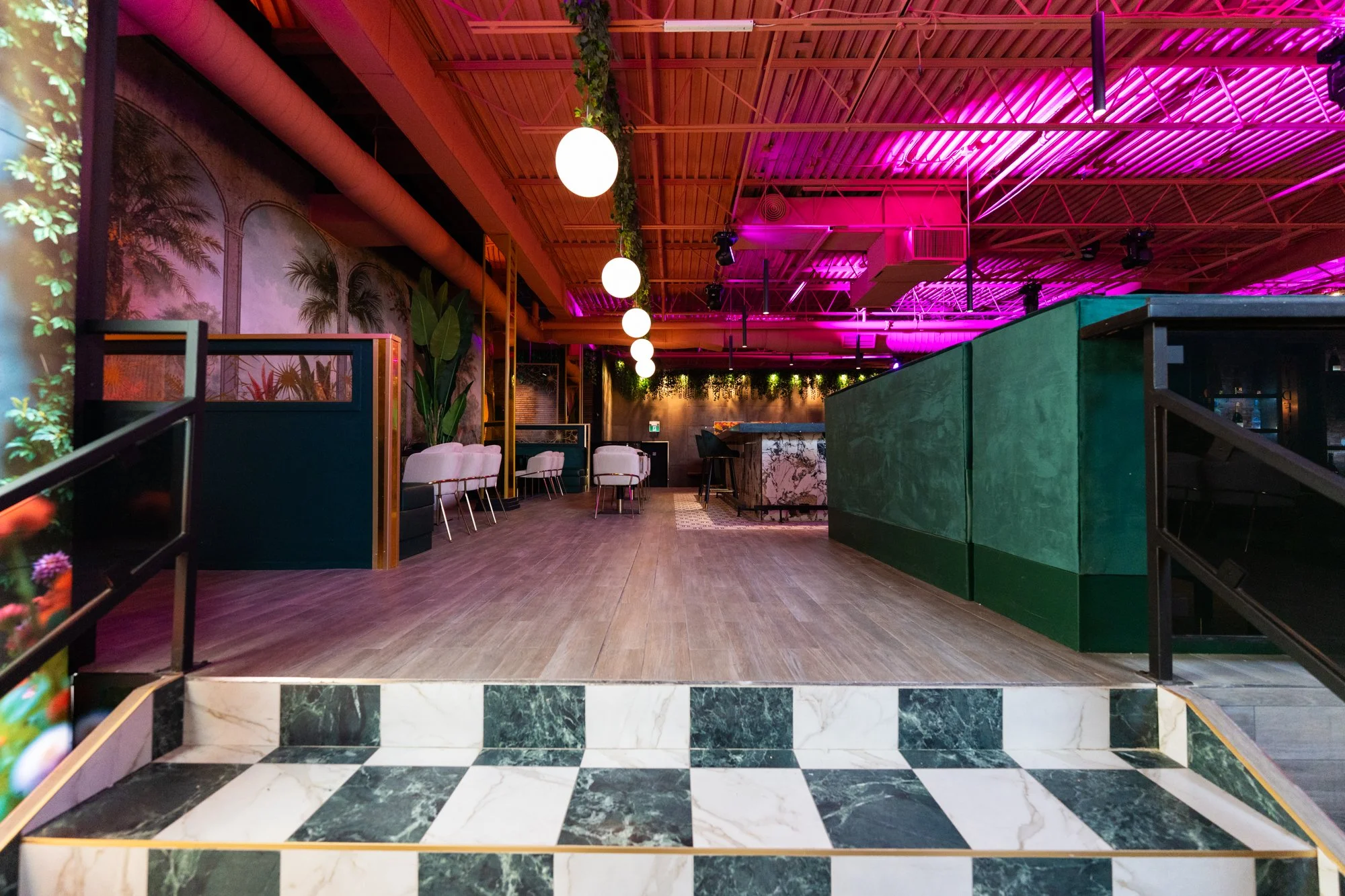 Empty nightclub with a small stage, tropical plants, lounge chairs, marble bar, colorful lighting, and a checkered marble floor.