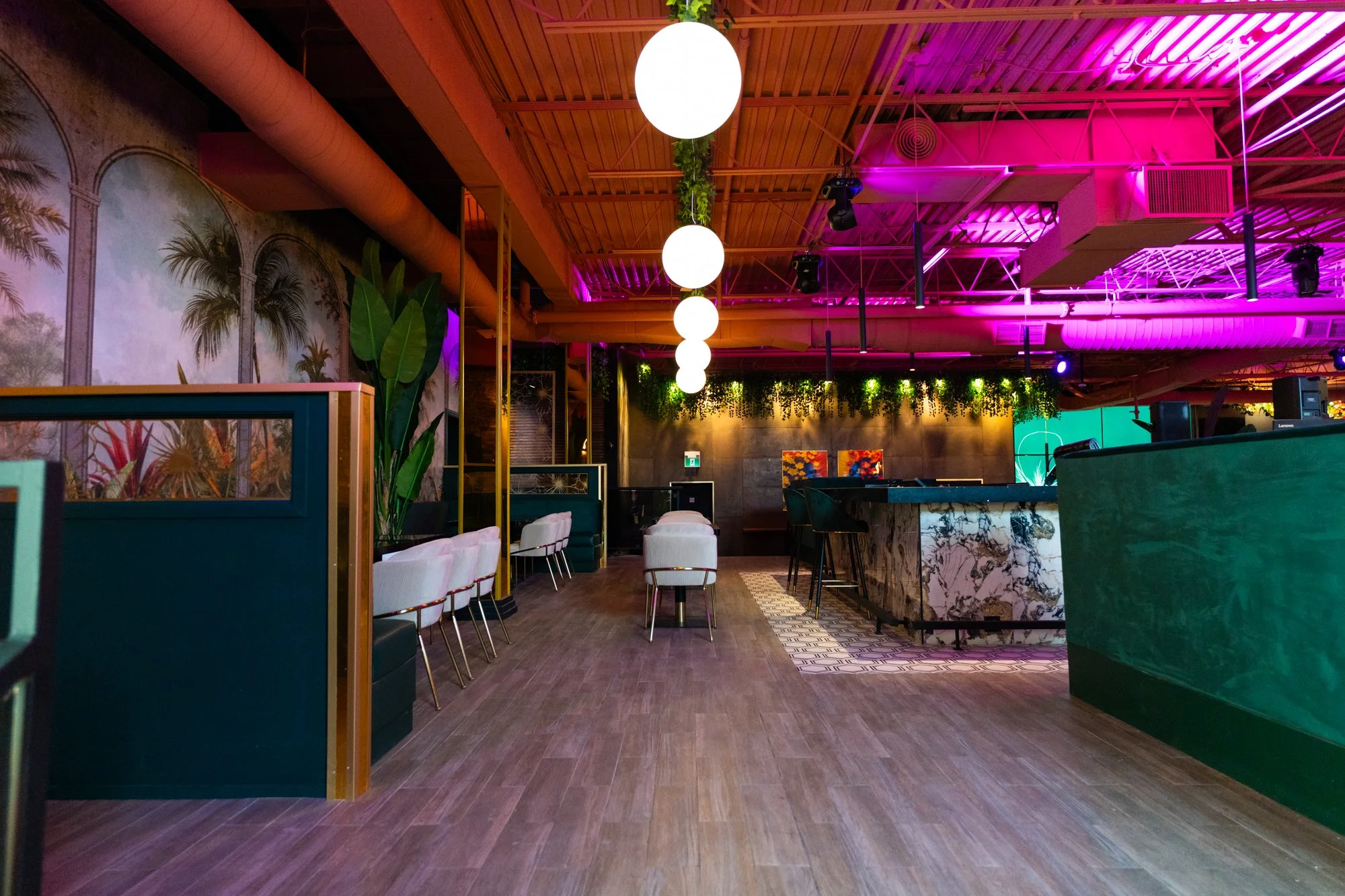 Interior of a modern bar or lounge with hanging white spherical lights, colorful neon lighting, tropical artwork on a wall, large green plants, a marble-topped bar with barstools, and seating areas with chairs and sofas.