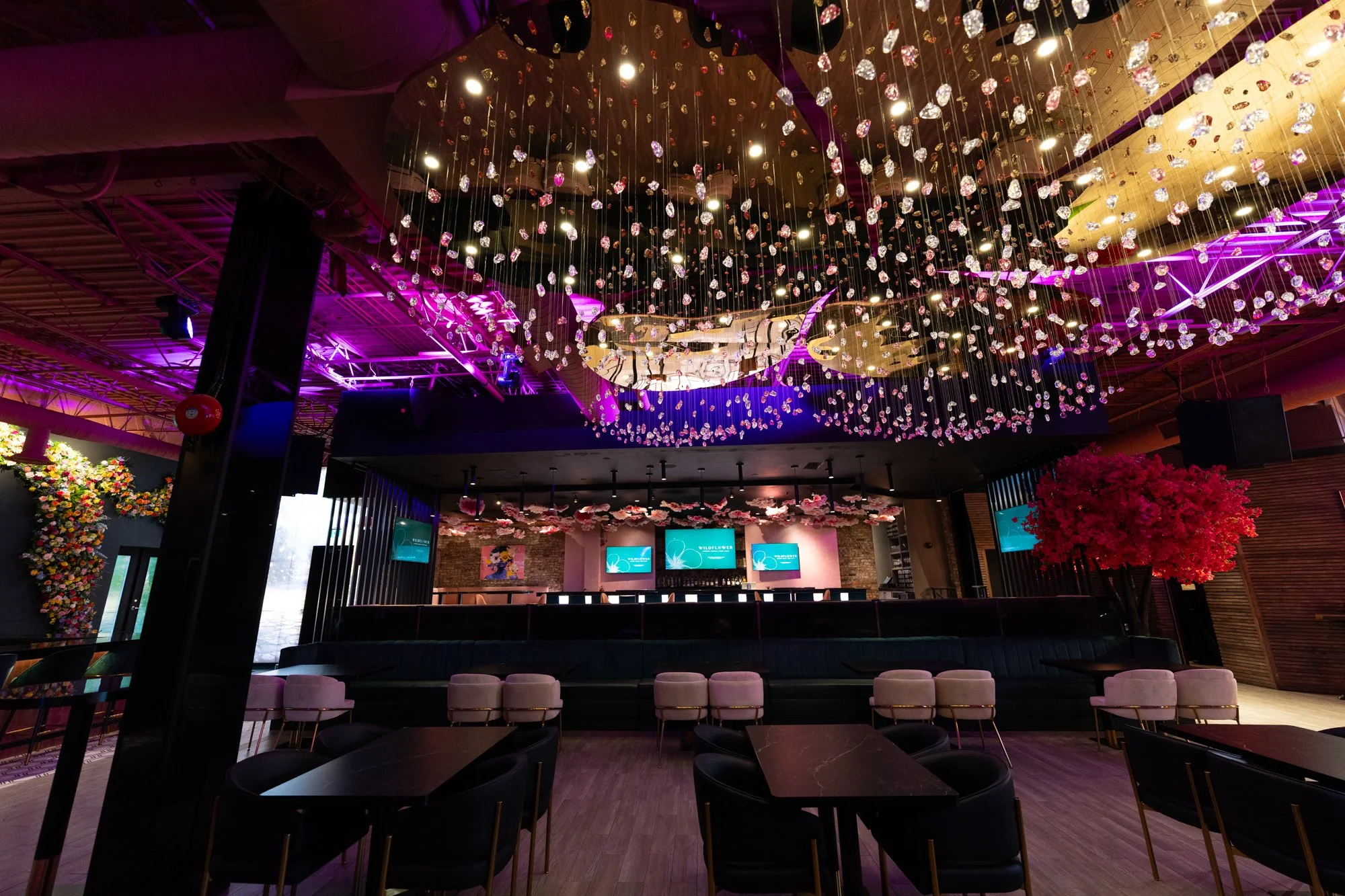 Interior of a modern event space or restaurant with a ceiling decorated with hanging flower arrangements, pink and purple lighting, a large chandelier-like installation with small hanging lights and crystals, a bar or stage area with three screens, a