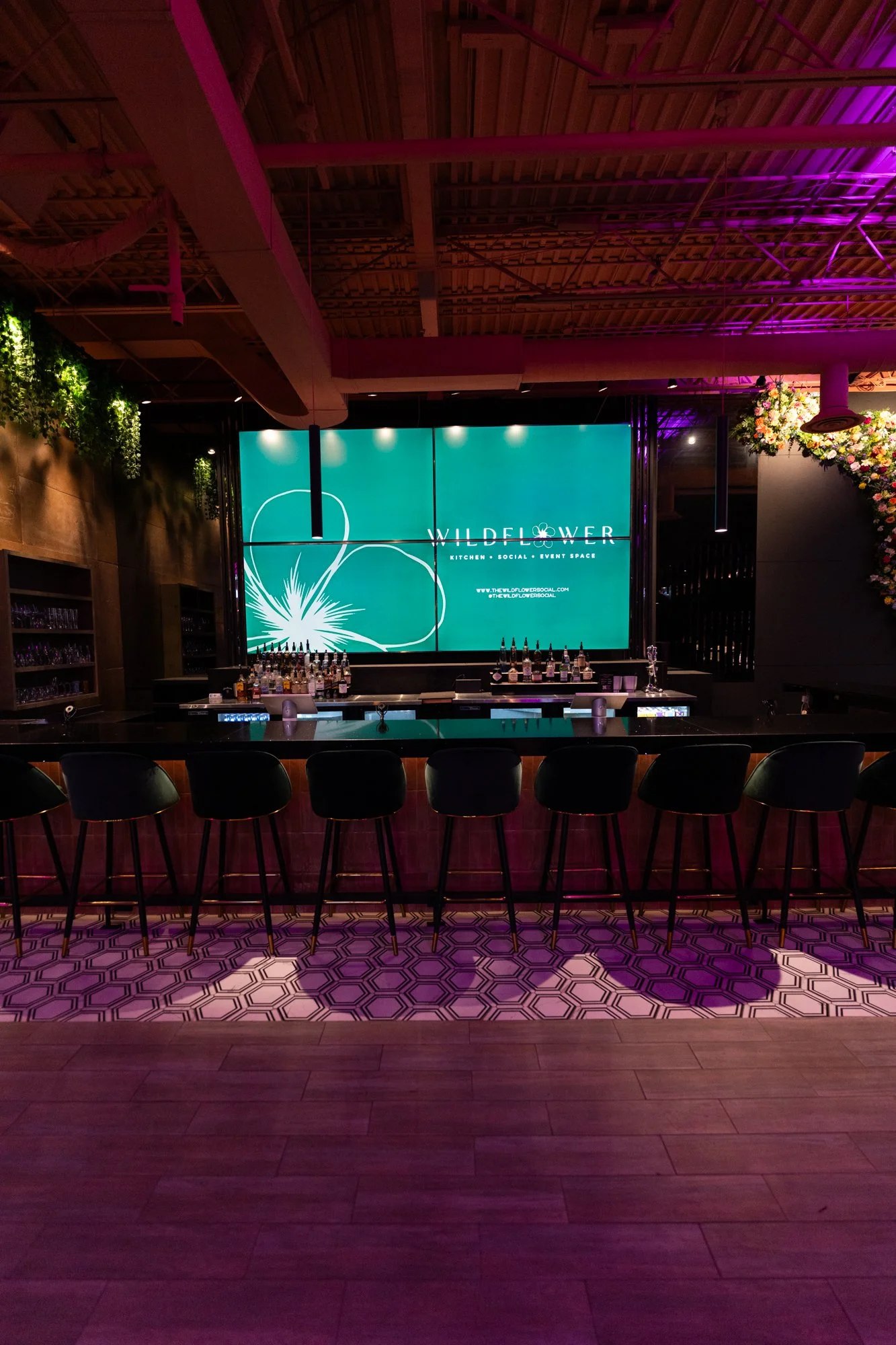 Indoor bar area at Wildflower social event space with a large digital display screen, bar counter with bottles, and black bar stools.