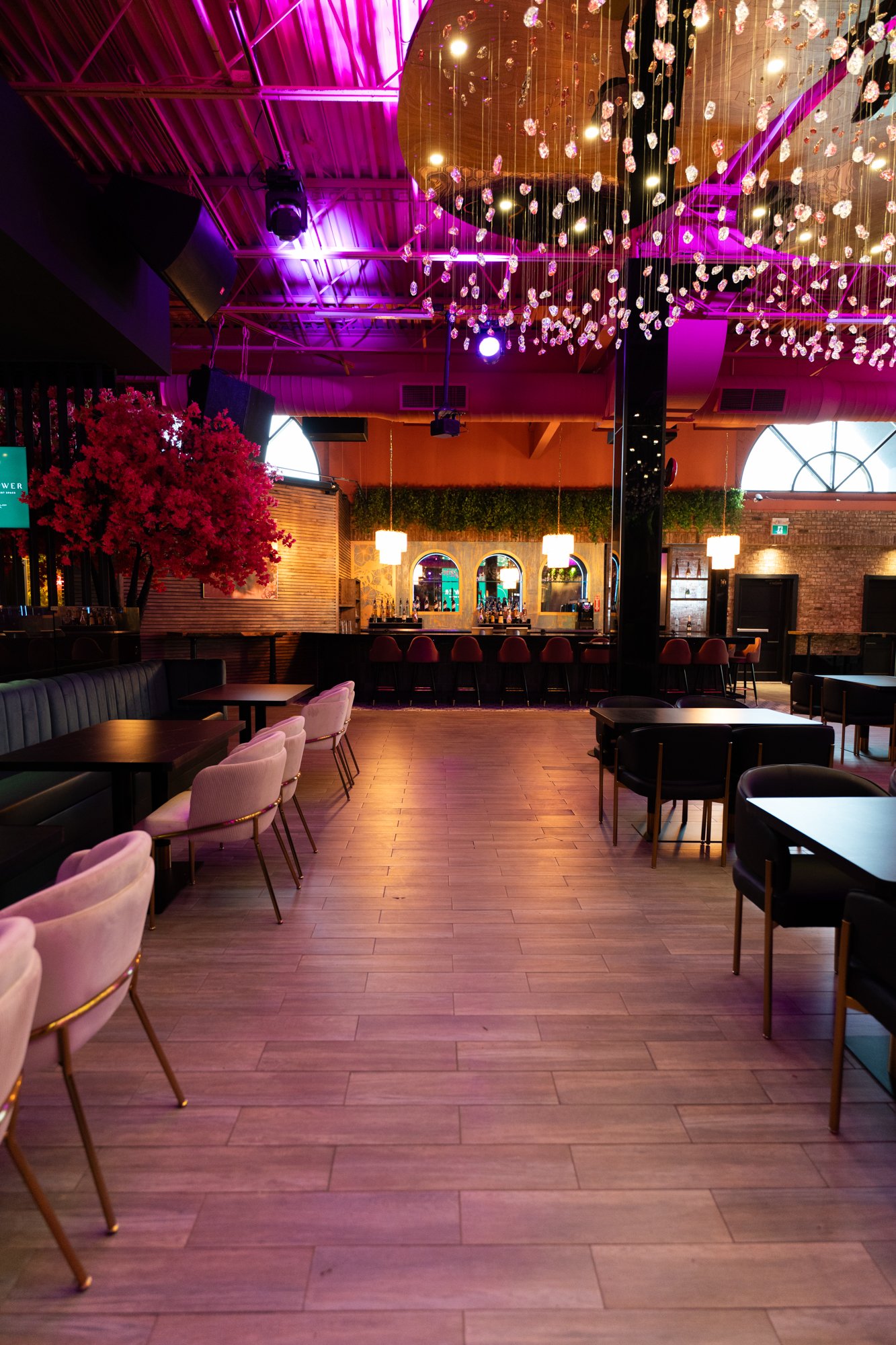 An empty modern bar and lounge with pink and purple lighting, wooden flooring, a bar counter with high stools, seating areas with chairs and tables, and decorative hanging crystals.