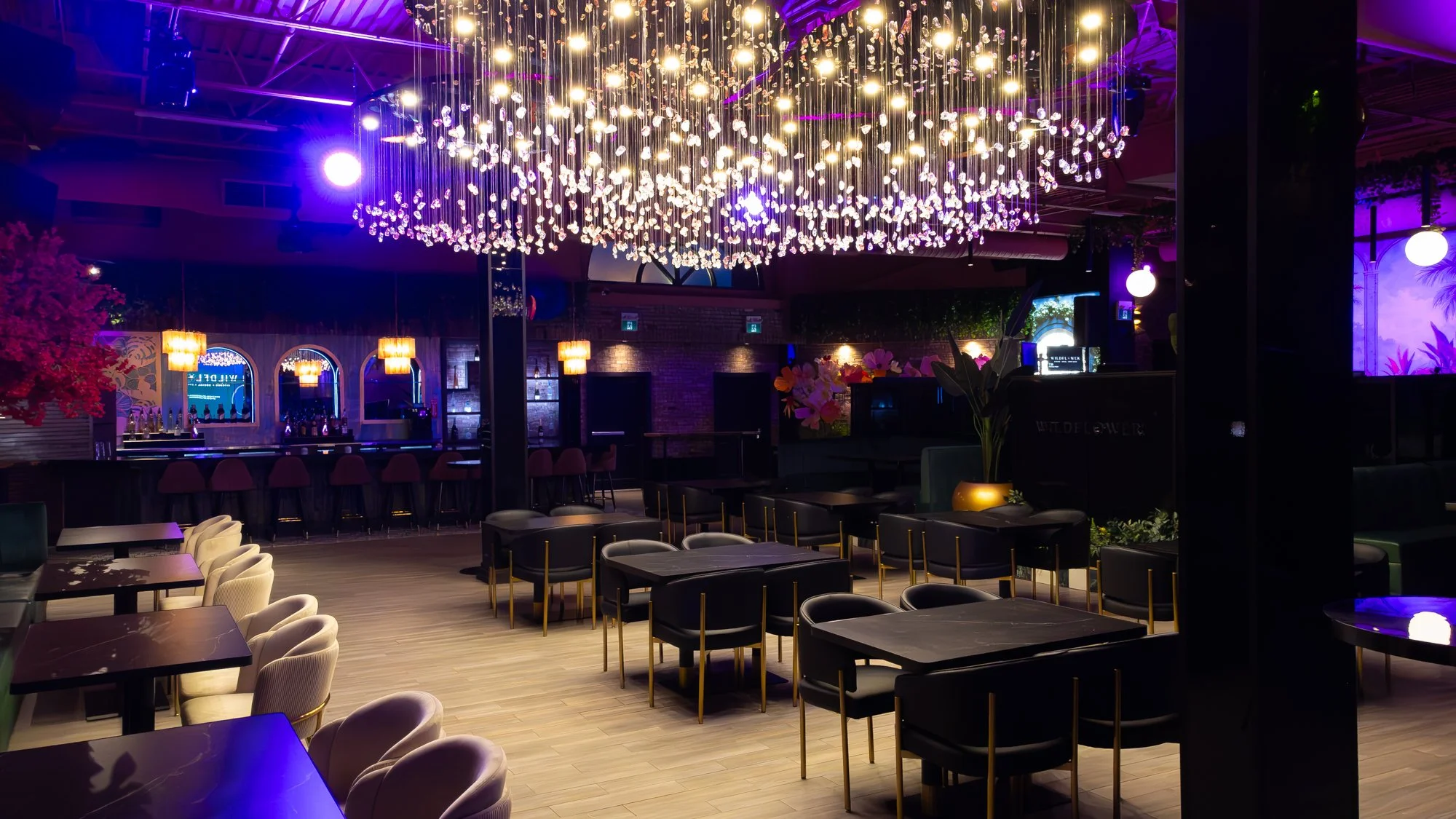 Interior of a modern bar or lounge with a large illuminated chandelier hanging from the ceiling, purple and warm lighting, a bar area with stools, and seating arrangements with tables and chairs.