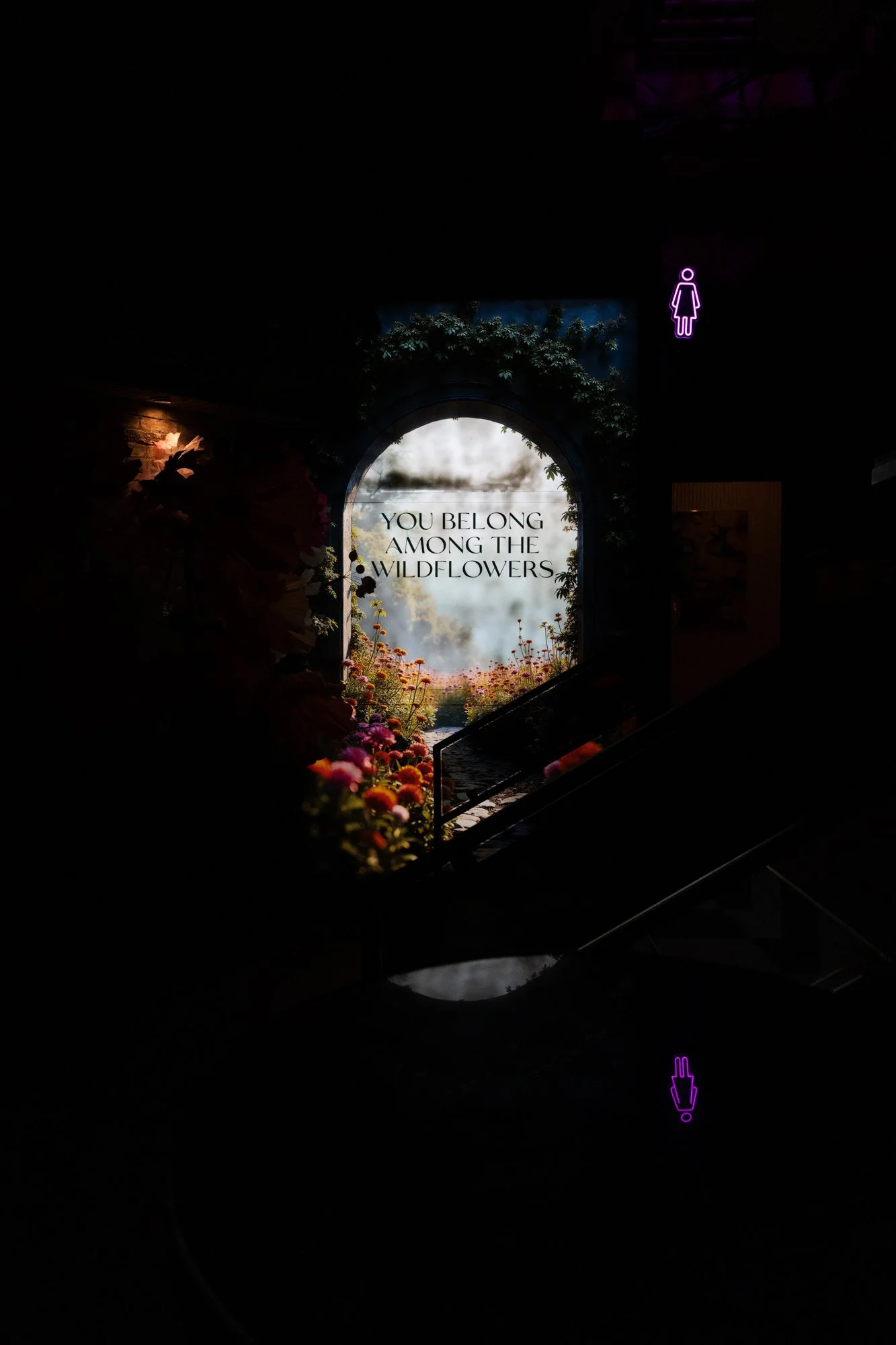 A dark indoor scene with a lit sign on a window that reads 'You belong among the wildflowers' surrounded by colorful flowers and greenery.
