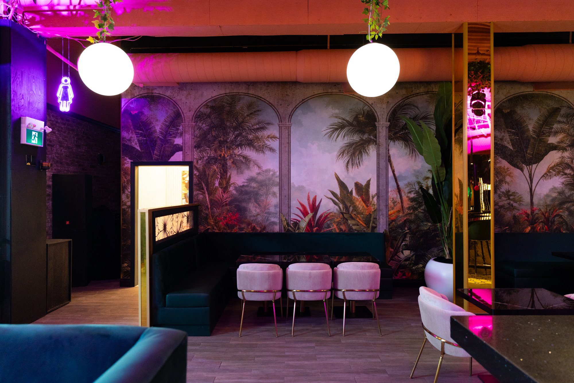 Interior of a modern lounge or bar with tropical-themed mural wallpaper, neon lighting, round pendant lights, and mixed seating including pink chairs and black booths.
