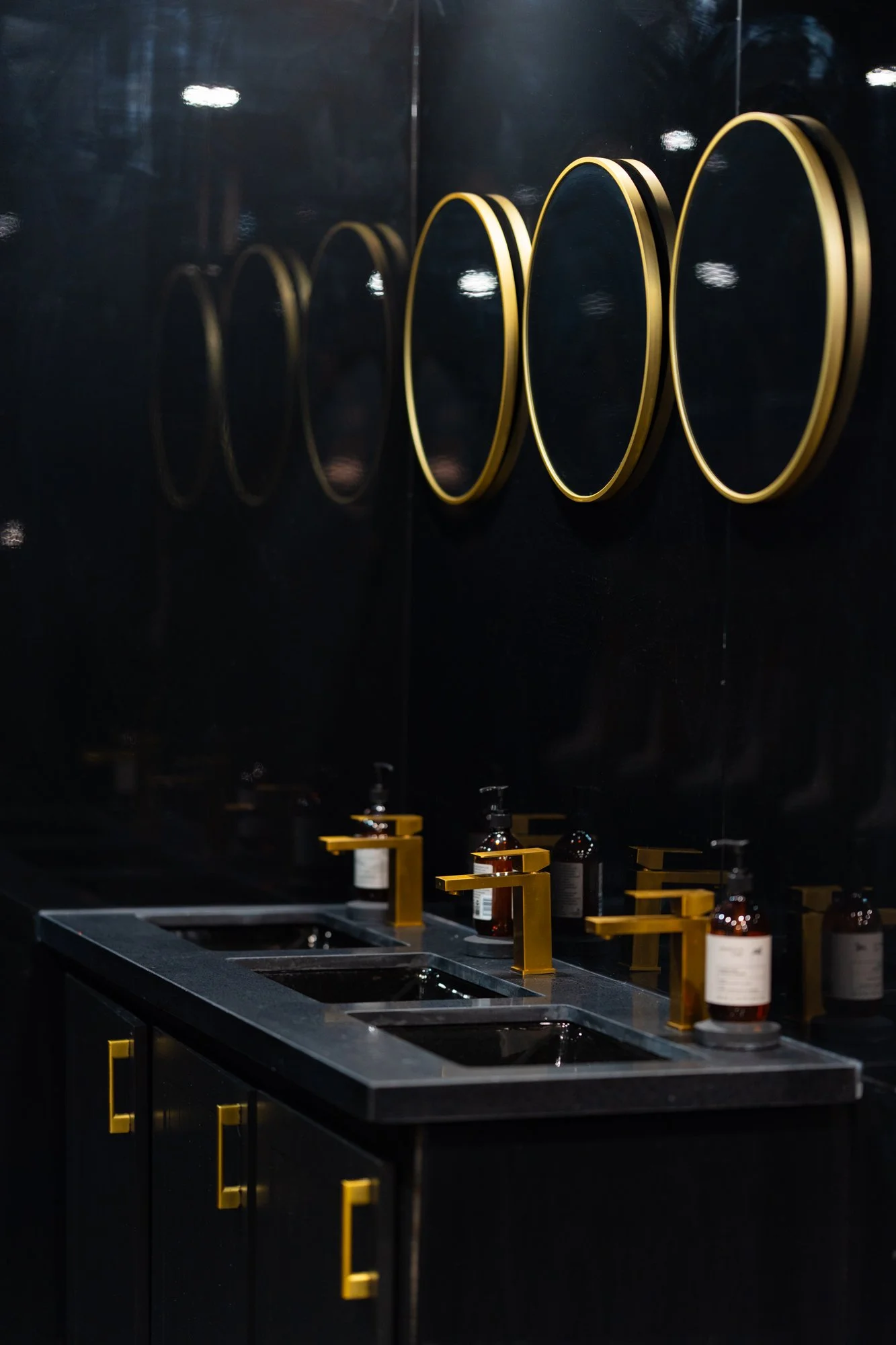 Black bathroom vanity with four sinks, gold faucets, black countertop, and mounted gold circular mirrors on a black wall. Bottles with pumps are placed on the countertop.