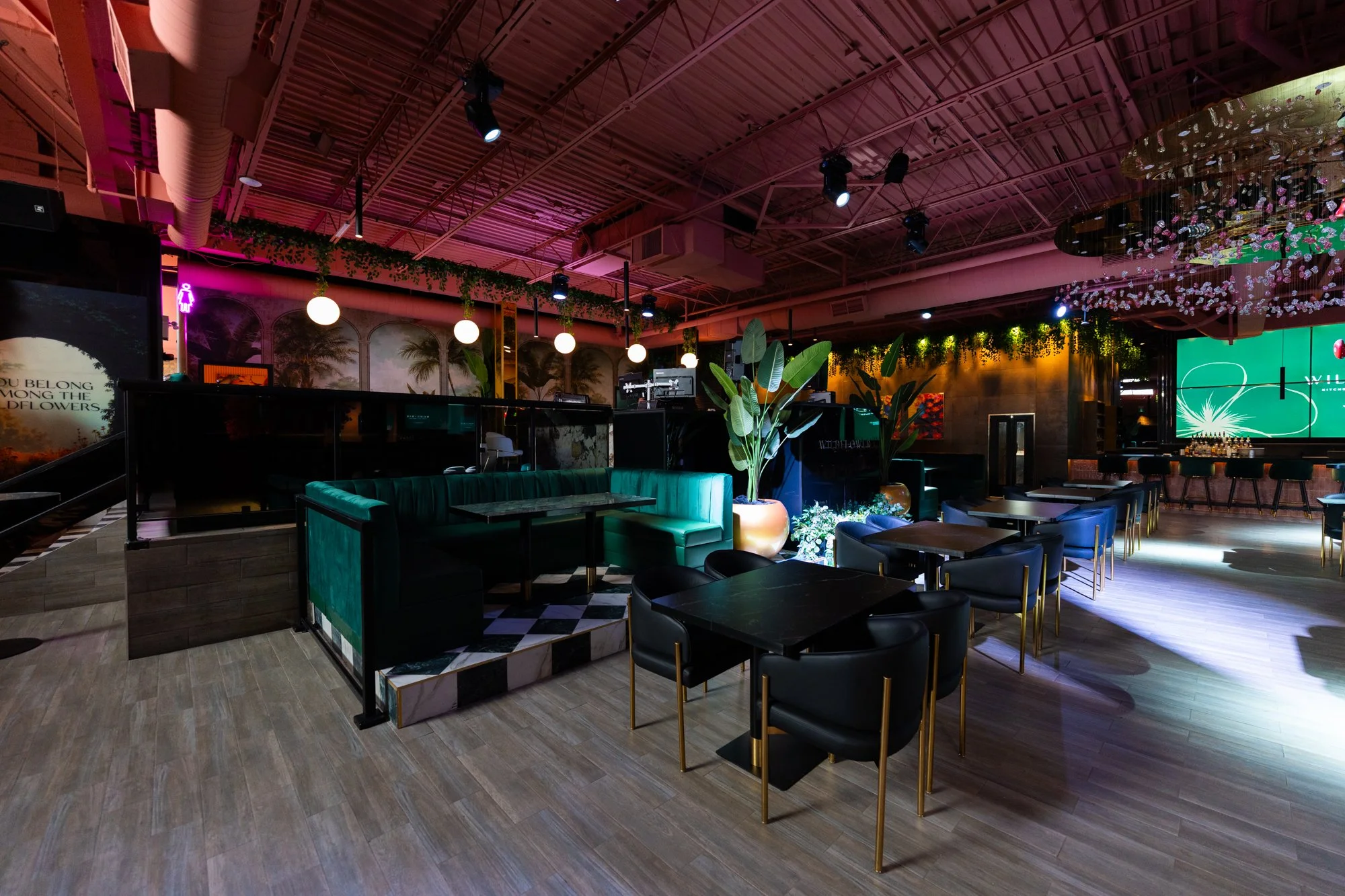 Interior of a nightclub or lounge with dim lighting, green and black seating, potted plants, and vibrant decor, including a bar area and large digital screens.