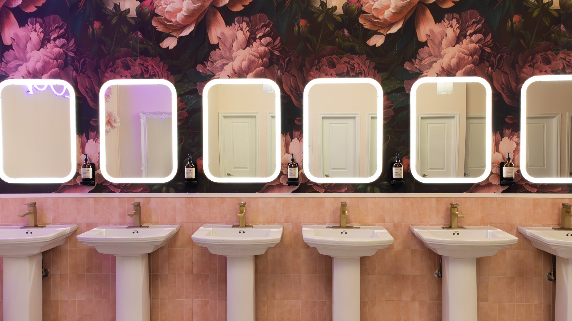 A row of five white sinks with gold faucets beneath illuminated rectangular mirrors on a floral wall in a bathroom or salon.