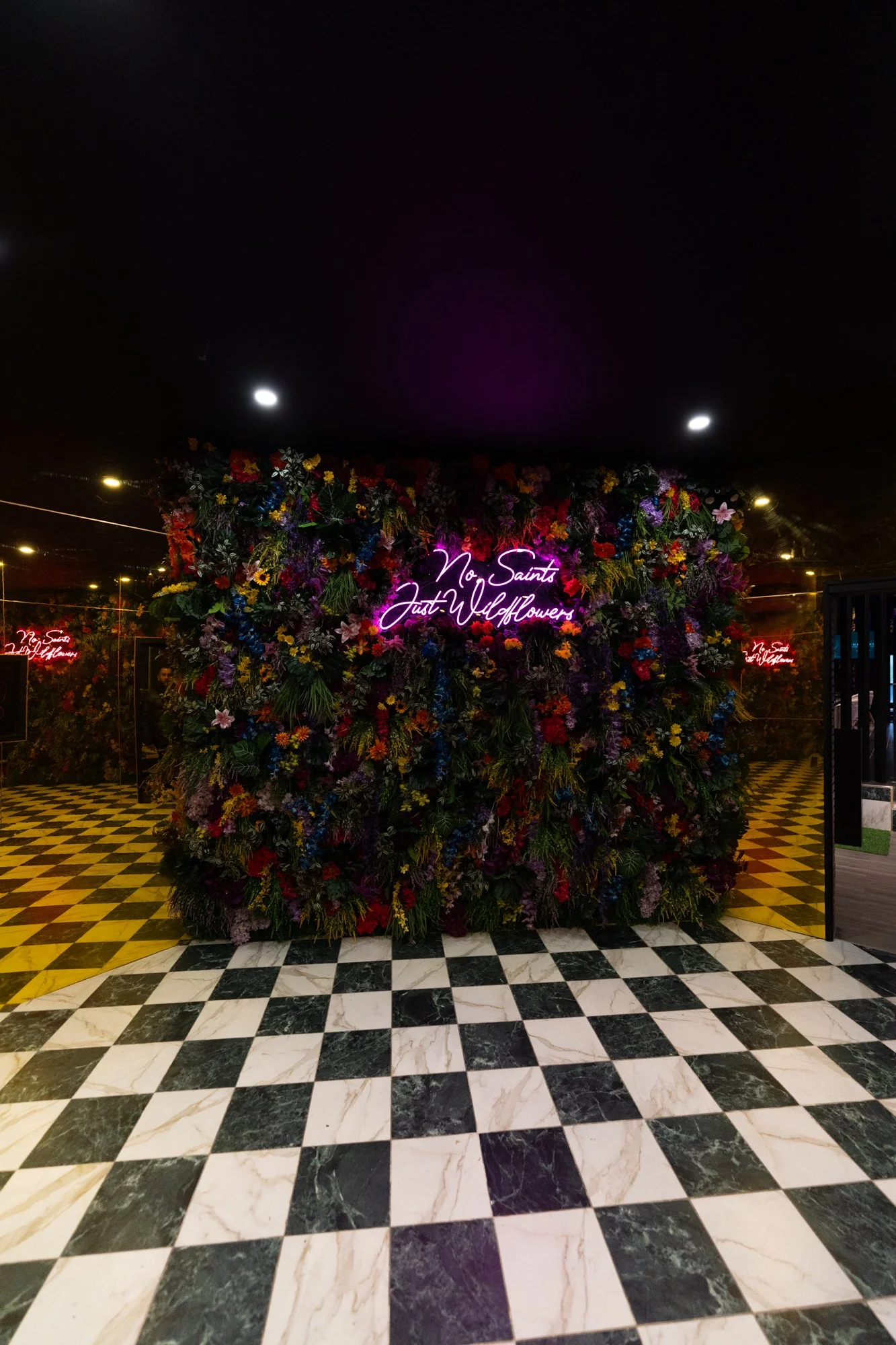 A floral wall with mixed colorful flowers and a neon sign that reads 'No Saint Just Wildflowers', set in a room with black and white checkered floor tiles.