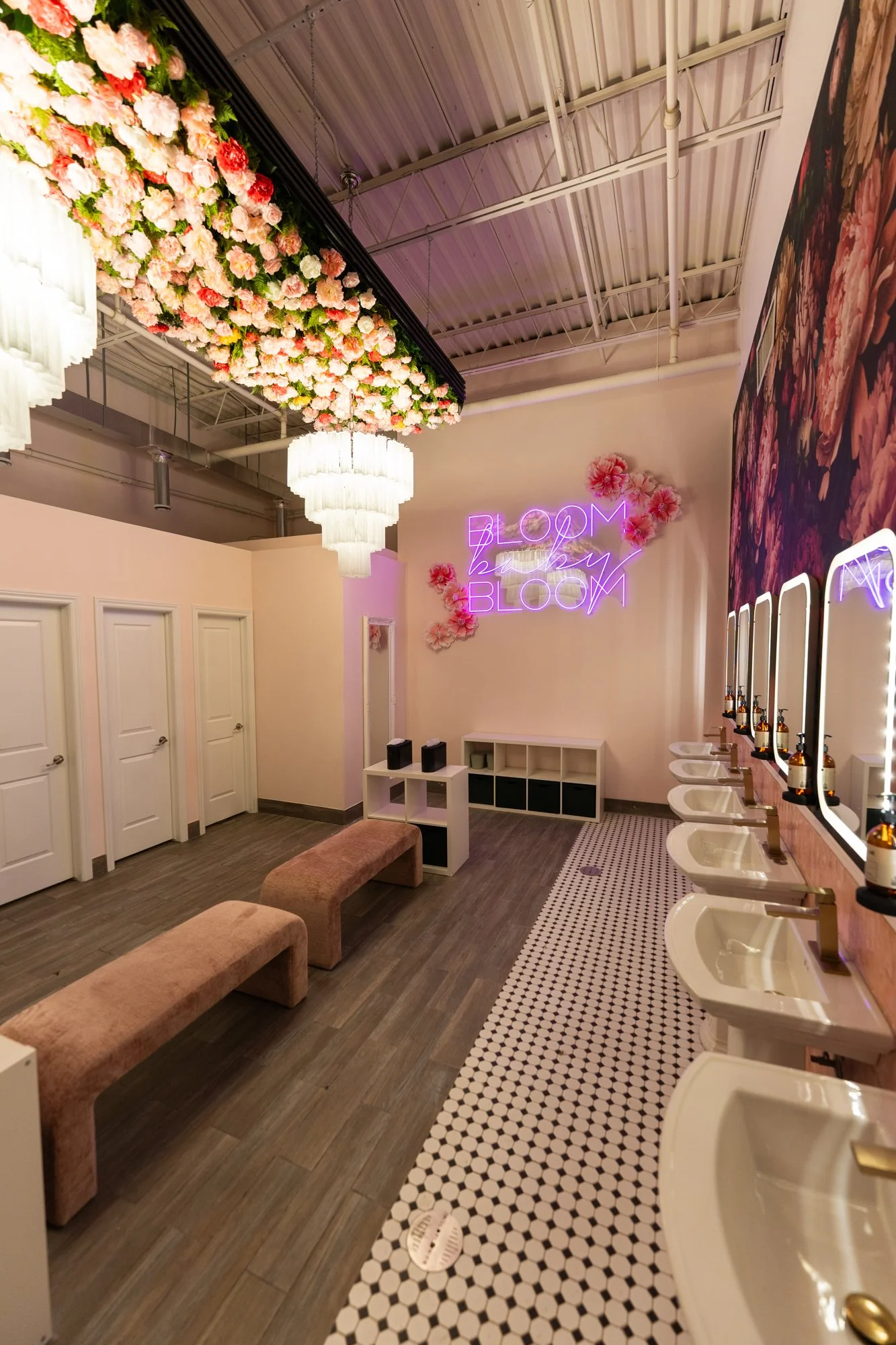 A bathroom decorated with pink and white flowers, including a flower canopy on the ceiling and a neon sign that says "BLOOM bloom" on the wall, along with a row of sinks and mirrors.