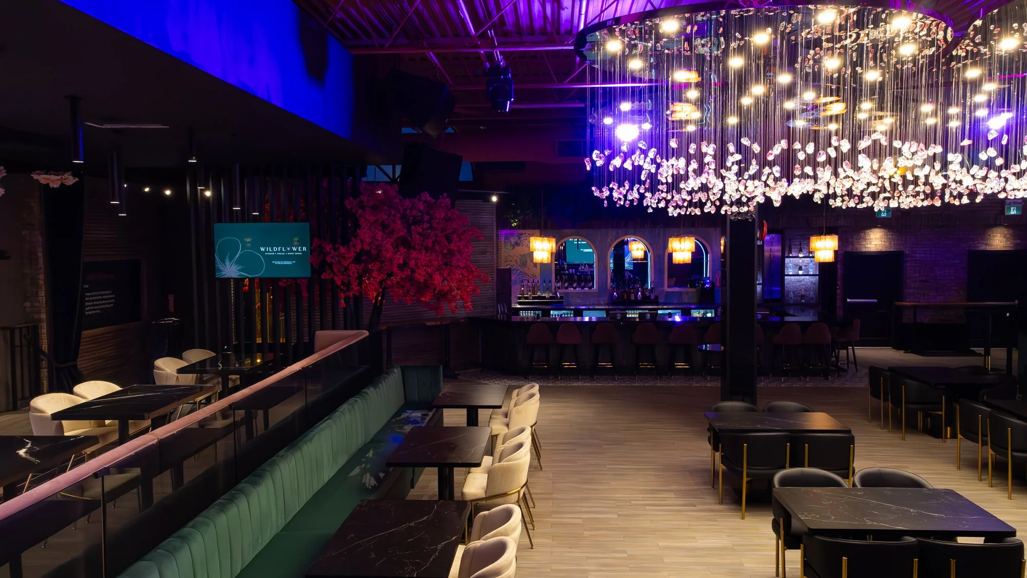 Interior of a modern bar or lounge with a large colorful flower chandelier, bar counter with stools, dining tables with chairs, a pink flowering cherry blossom tree, and purple accent lighting.