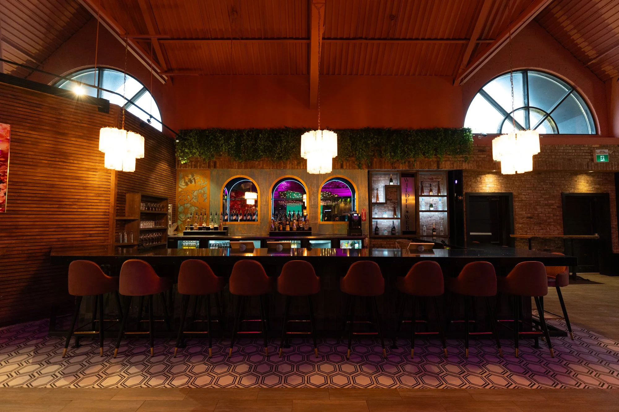 Interior of a modern bar with a long black counter, red chairs, hanging white pendant lights, brick and wooden walls, arched windows, and colorful lighting in the background.