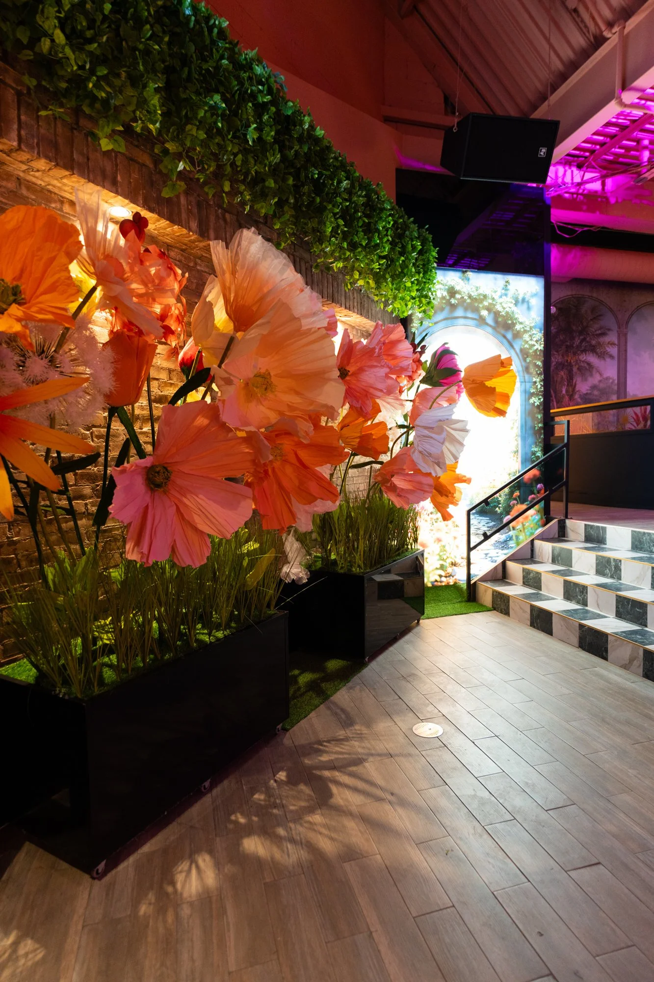 Indoor setting with large pink and orange artificial flowers in black planters, a brick wall, green foliage above, a lit backdrop, and marble stairs with a black railing.