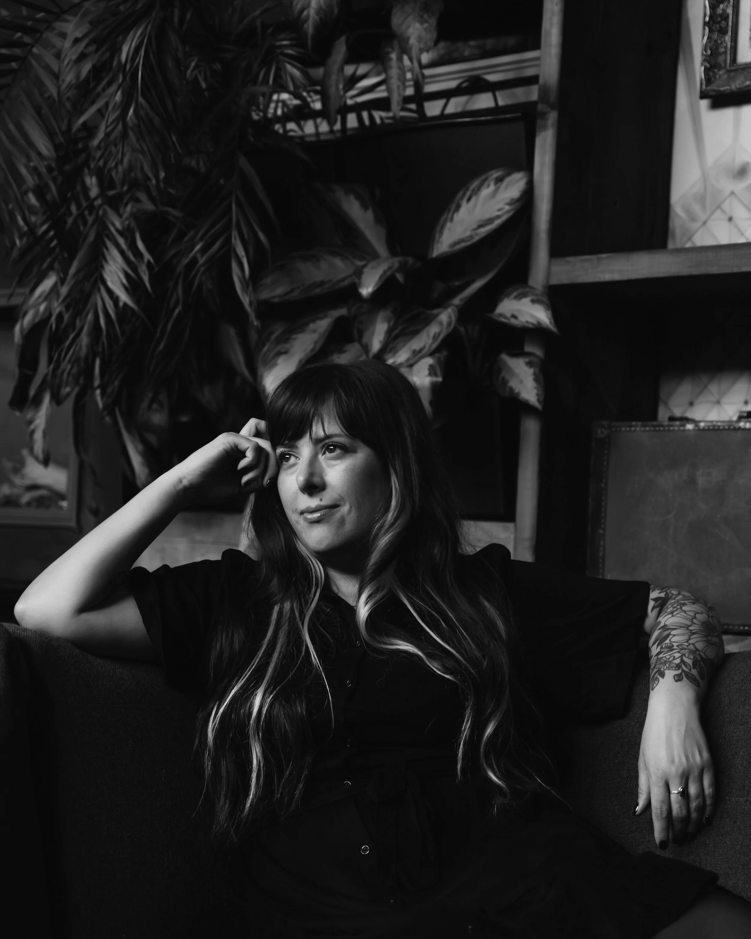 A black and white photograph of K.L. Speer showing a woman with long hair resting her head on her hand and sitting in front of leafy plants and furniture in a room.