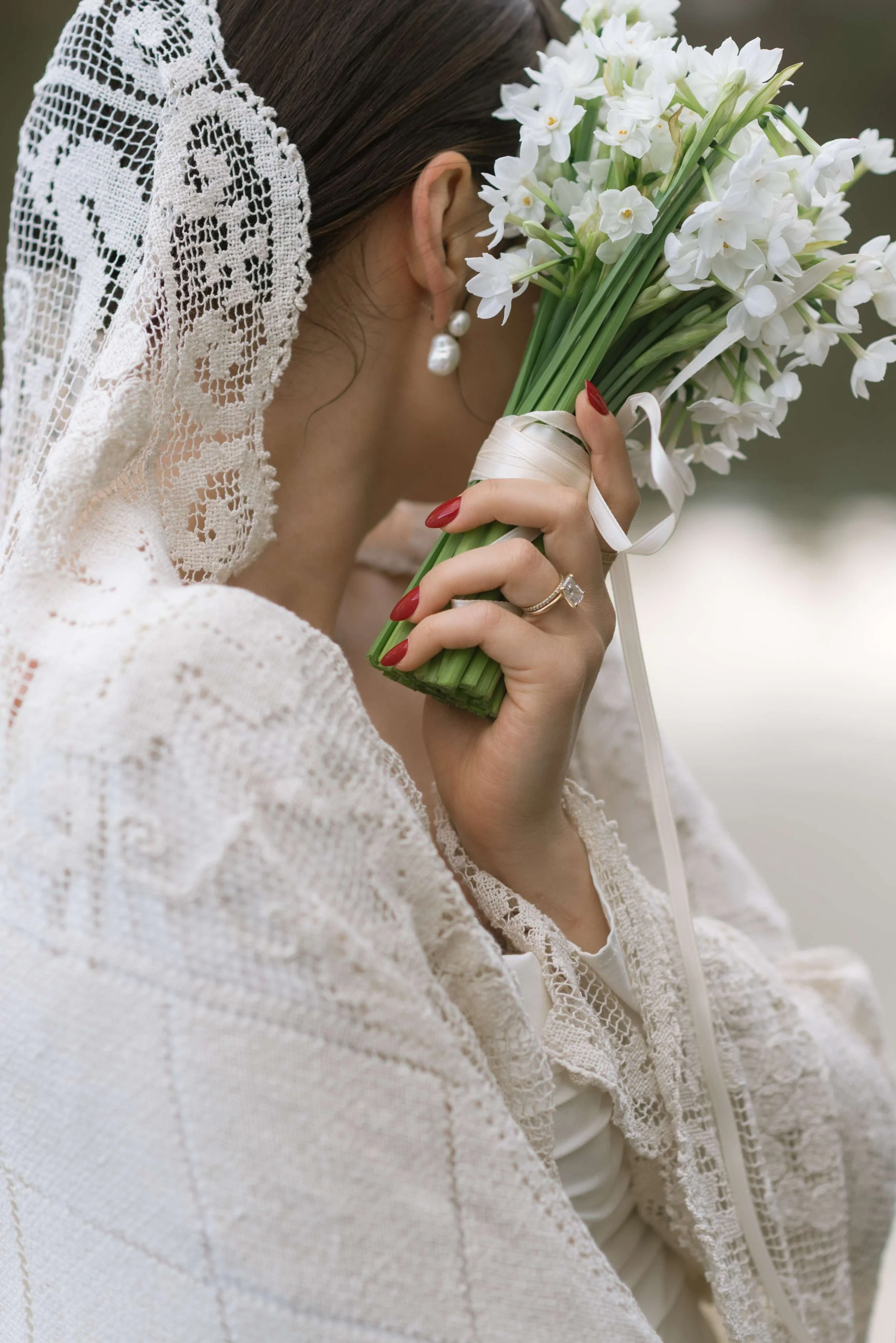 A woman dressed in vintage lace attire holds a bouquet of white flowers close to her face, wearing pearl earrings and a ring, with her dark hair partially covered by a lace head covering.