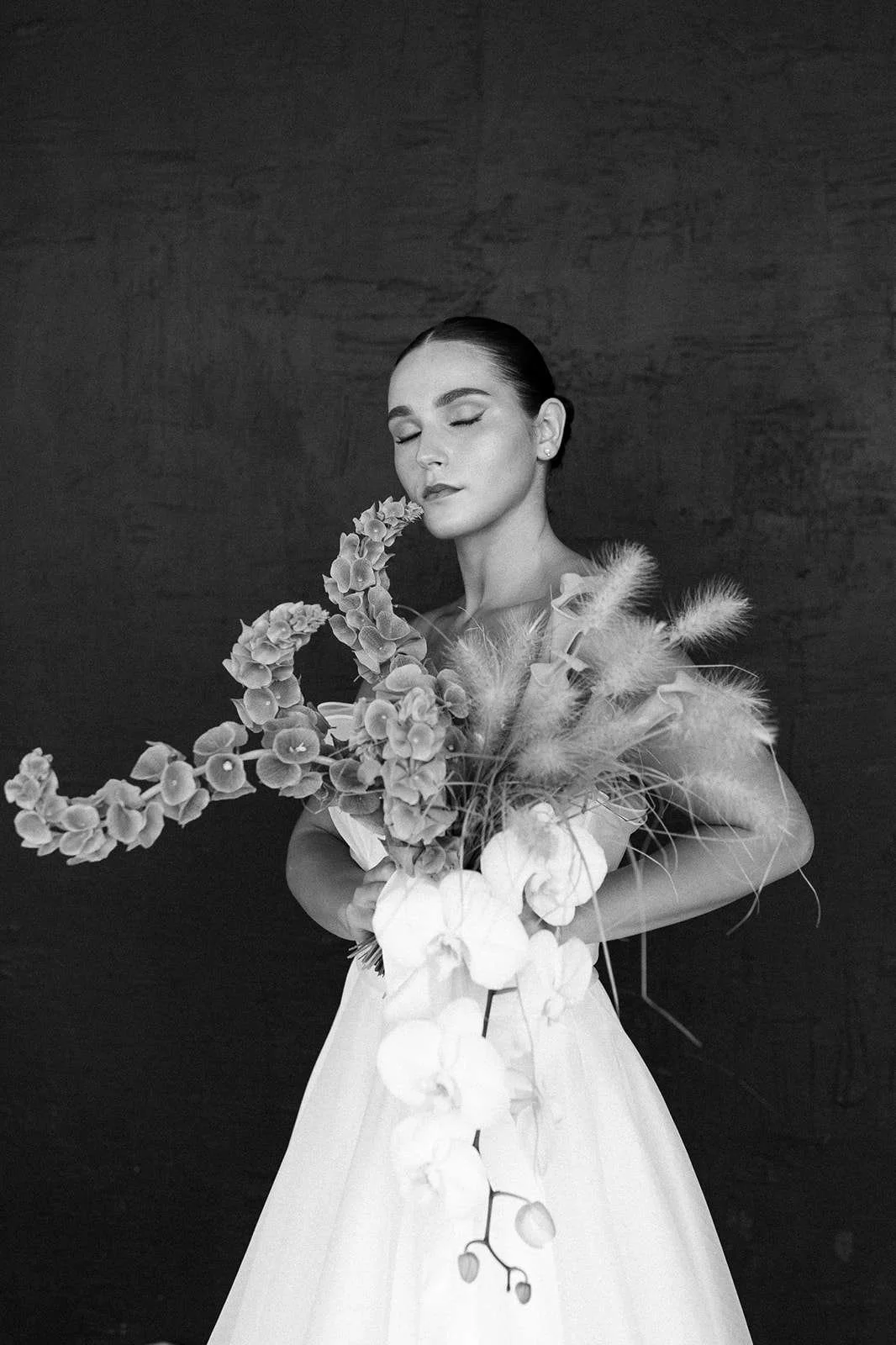 A woman with dark hair styled back and wearing a light-colored dress, holding a bouquet of large flowers and soft feathery foliage, with her eyes closed against a dark textured background.