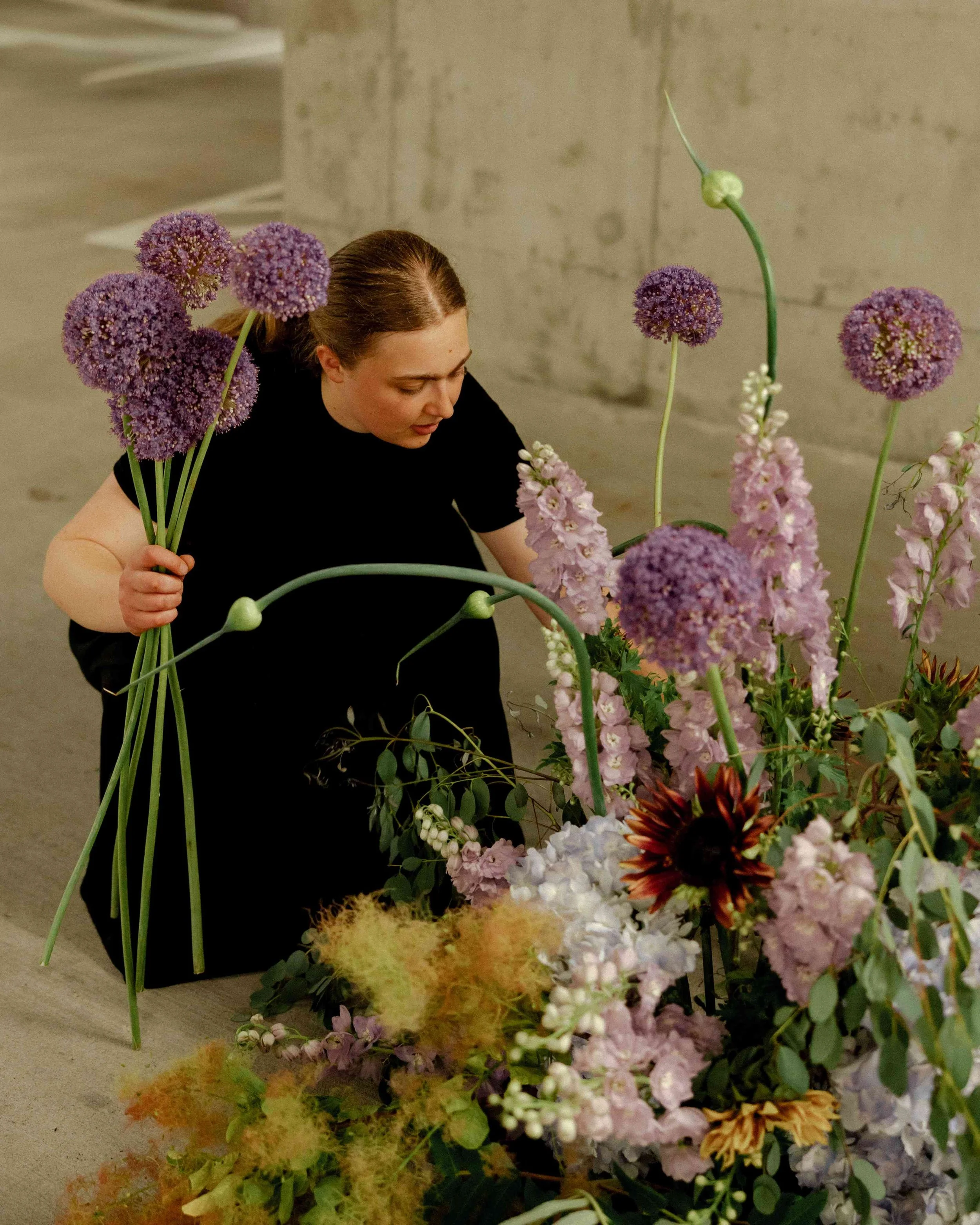 A woman arranging a colorful flower display with purple, pink, white, and orange flowers on a beige floor near a concrete wall.