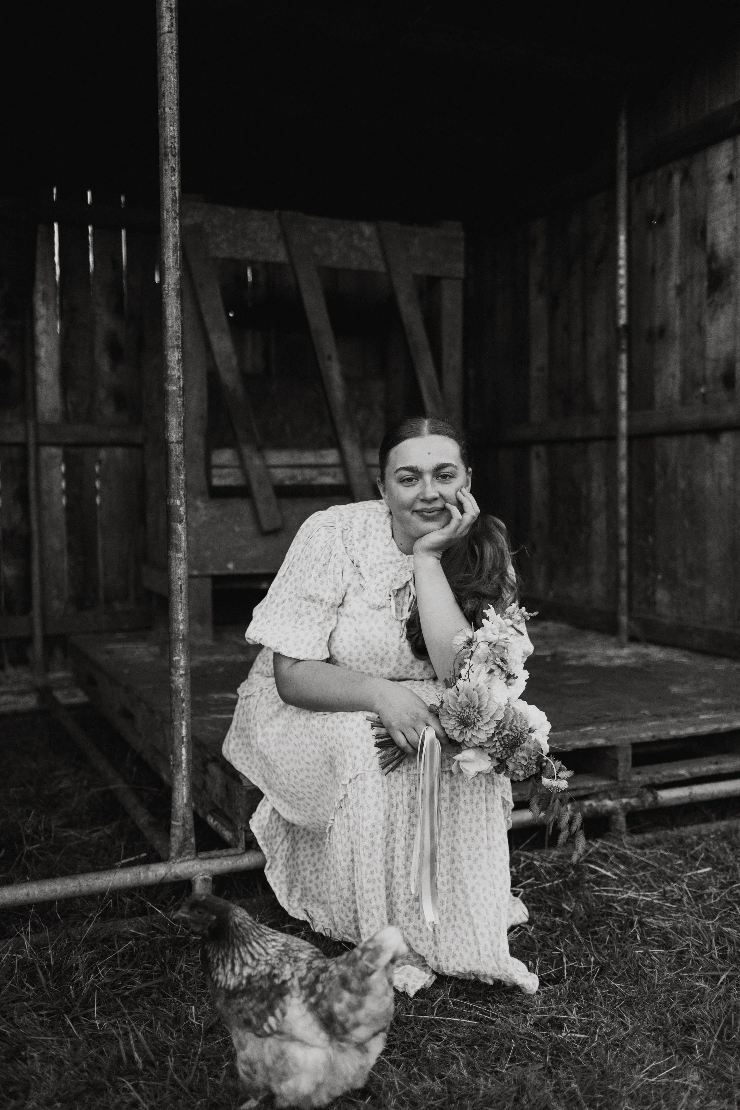 A woman in a light-colored dress sitting on the ground, holding a bouquet of flowers, while a hen stands nearby, in front of a wooden barn or shed.