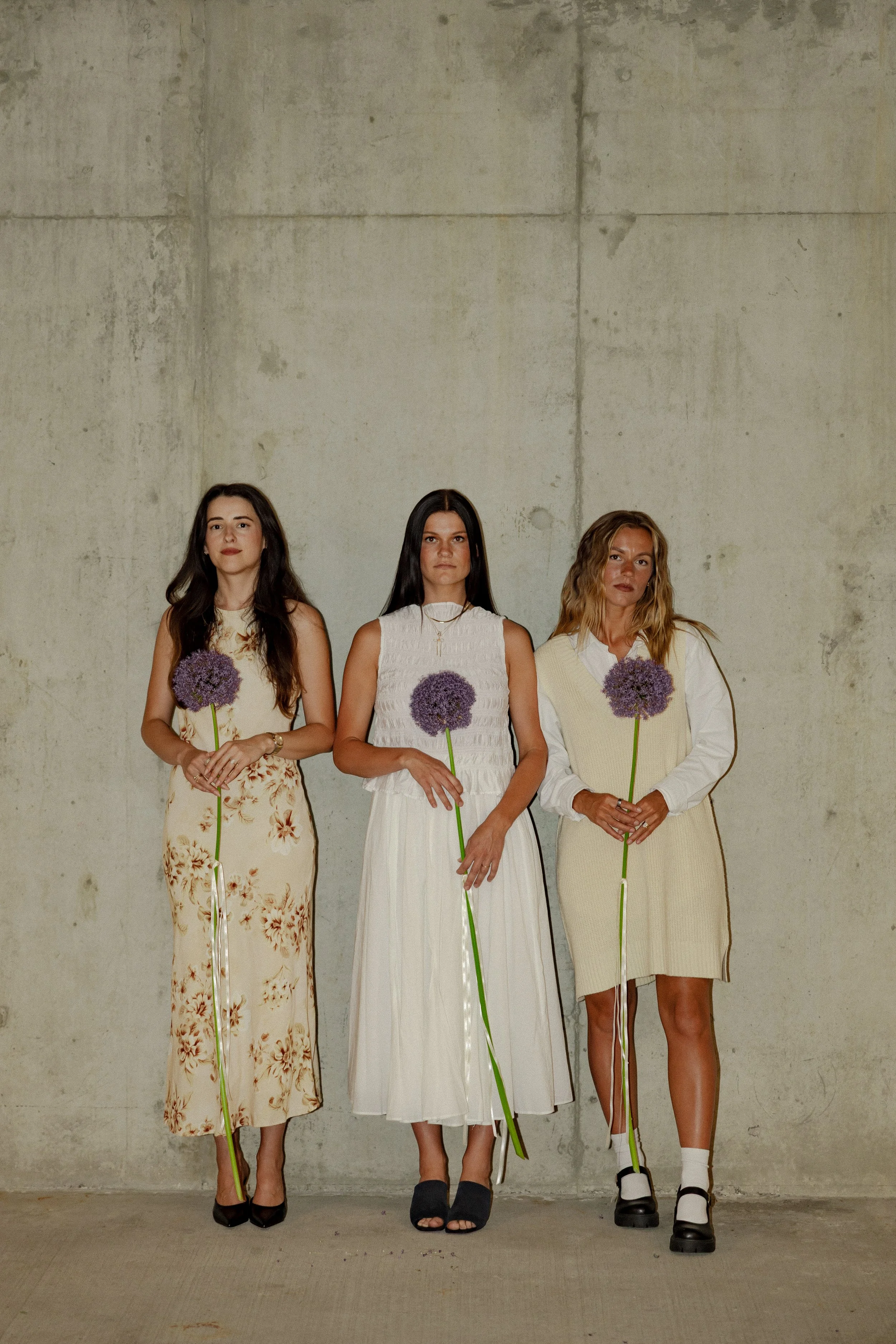 Three women standing side by side against a concrete wall, each holding a purple flower on a long green stem, dressed in stylish outfits with serious expressions.