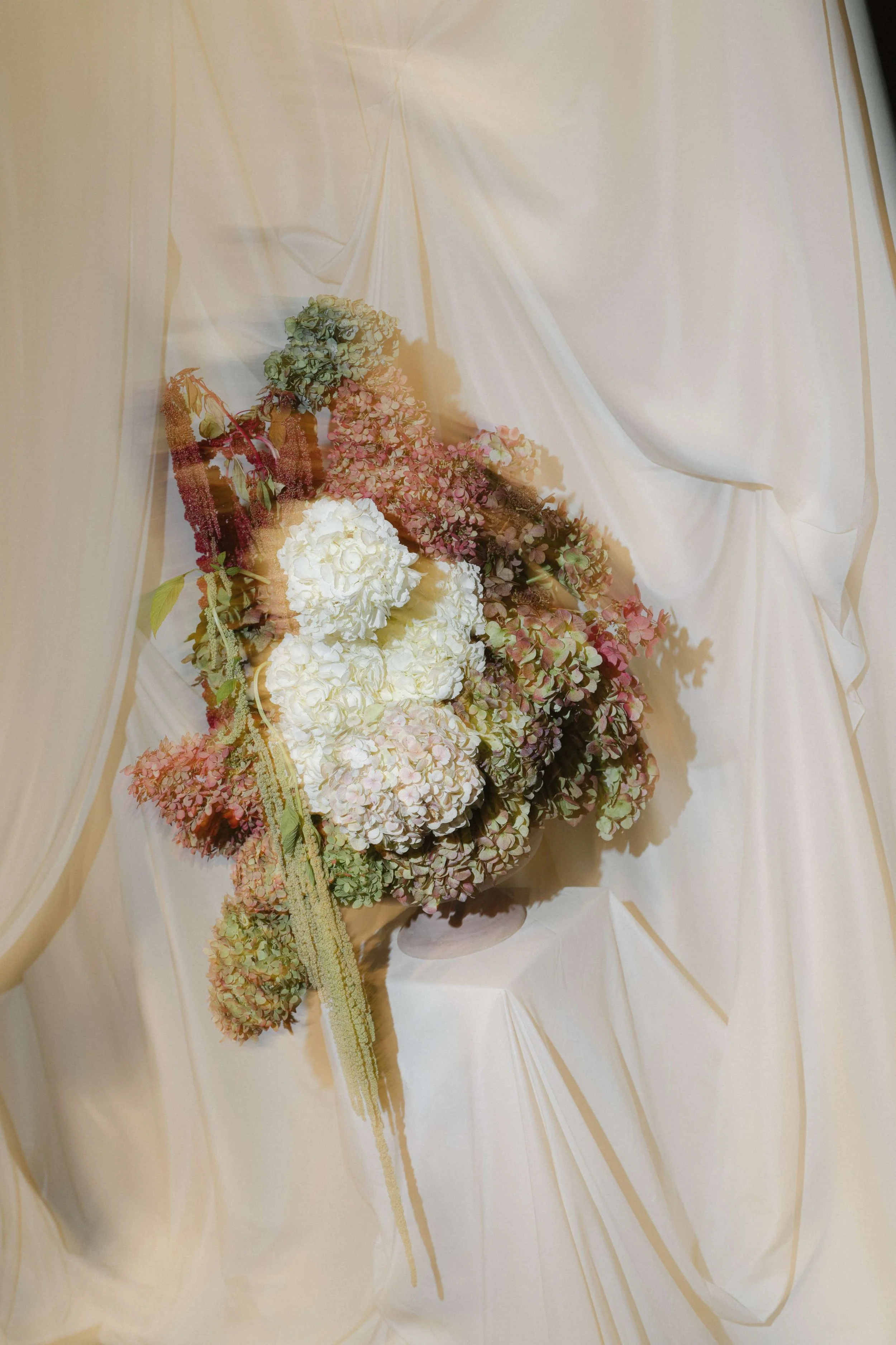 A floral arrangement consisting of pink, white, and green hydrangeas on a fabric backdrop.