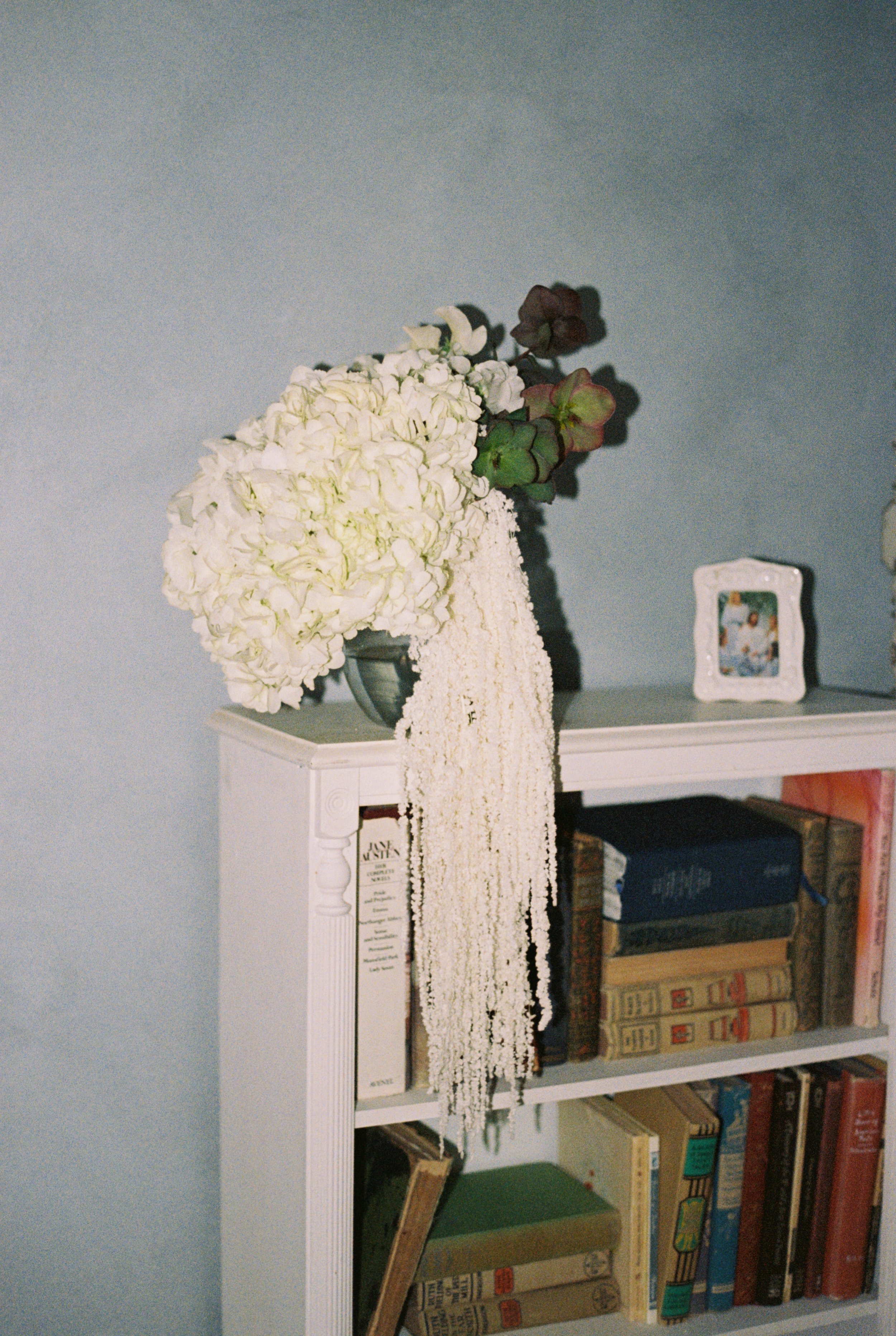 A white bookshelf filled with various books, a white floral arrangement on top, and a small picture frame on the right side.