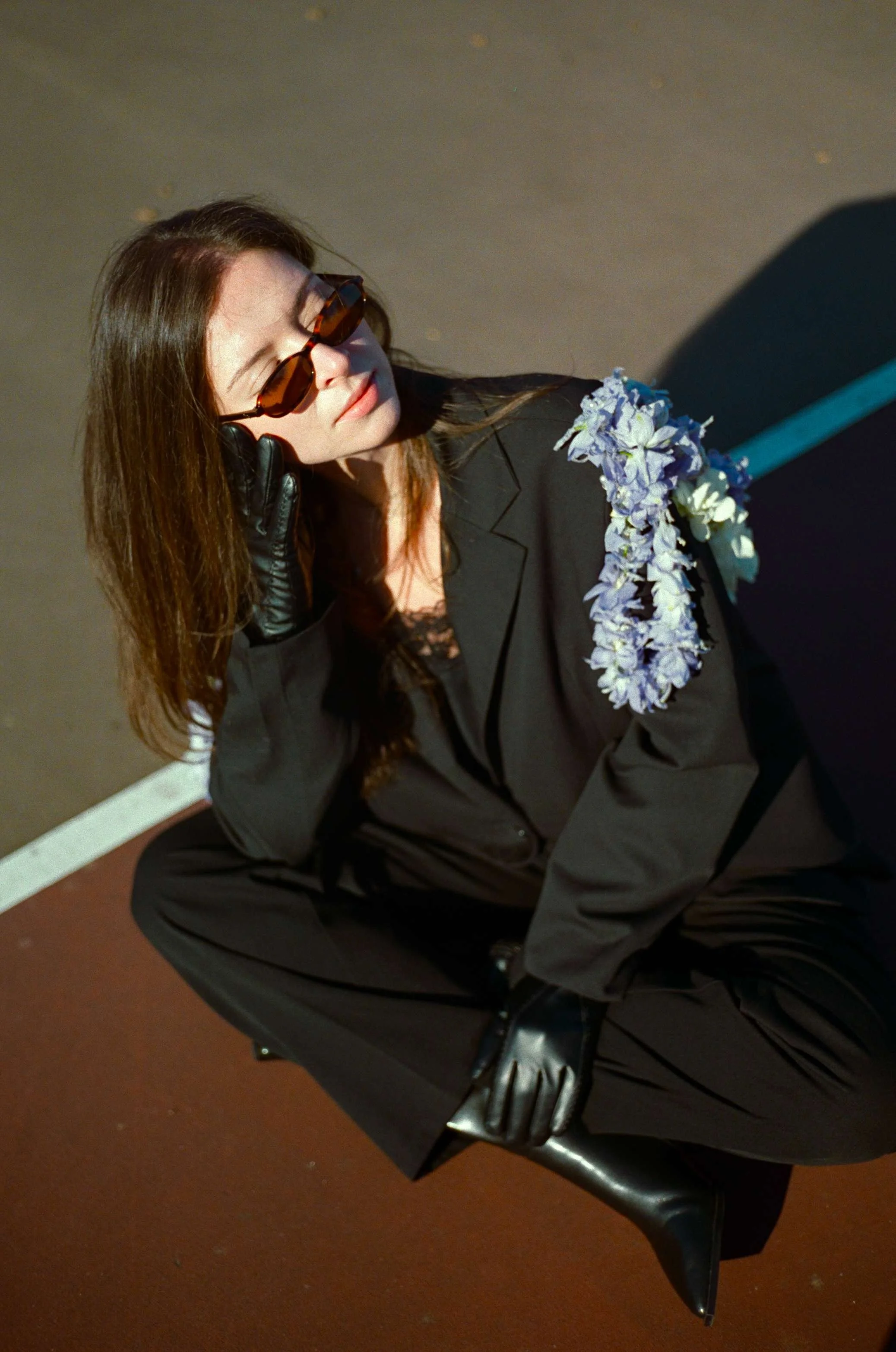 A woman with long brown hair and sunglasses, dressed in a black blazer, is sitting on a black chair with a floral lei on her shoulder, on a dimly lit surface.