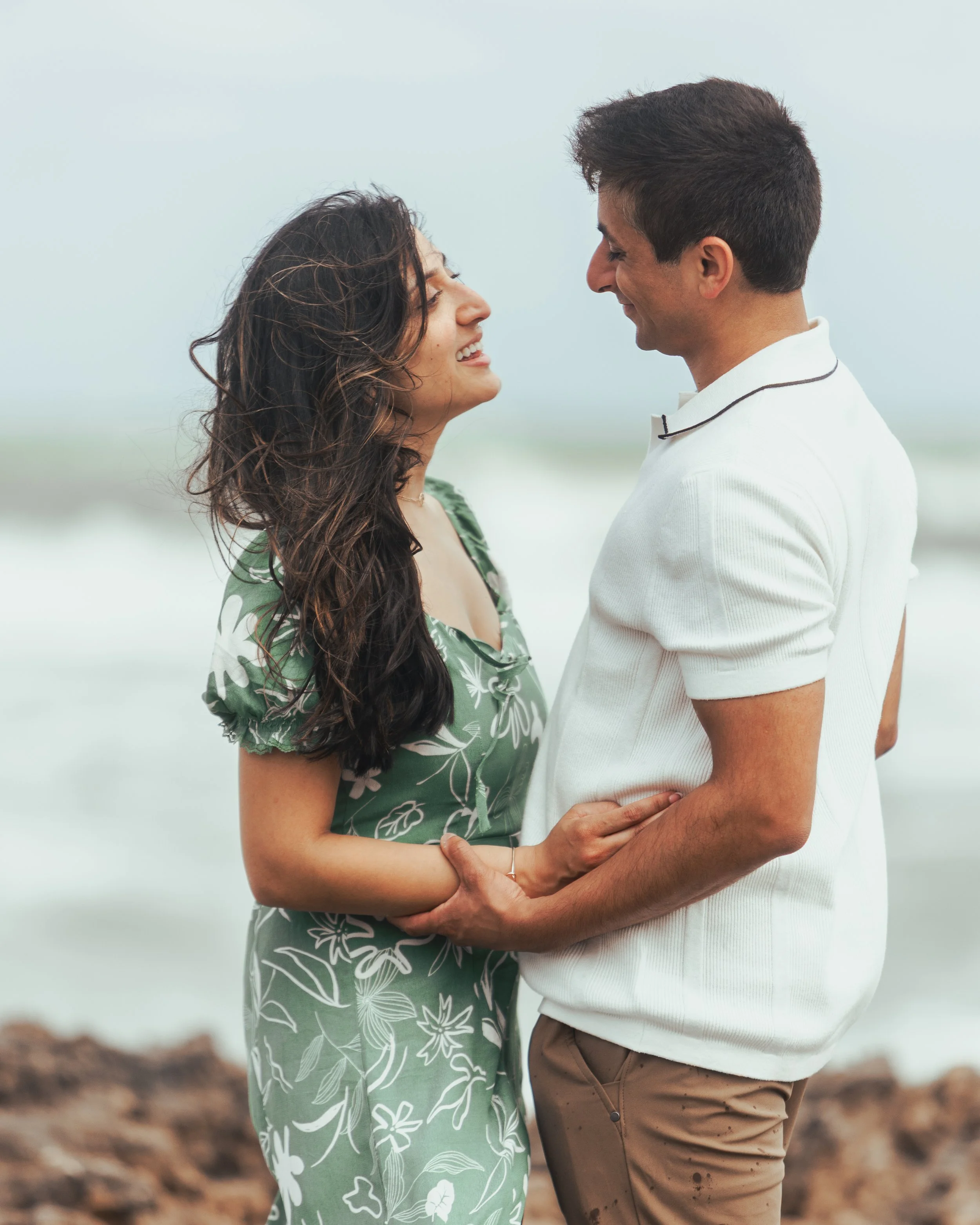 A smiling couple standing close together on a beach, looking into each other's eyes.