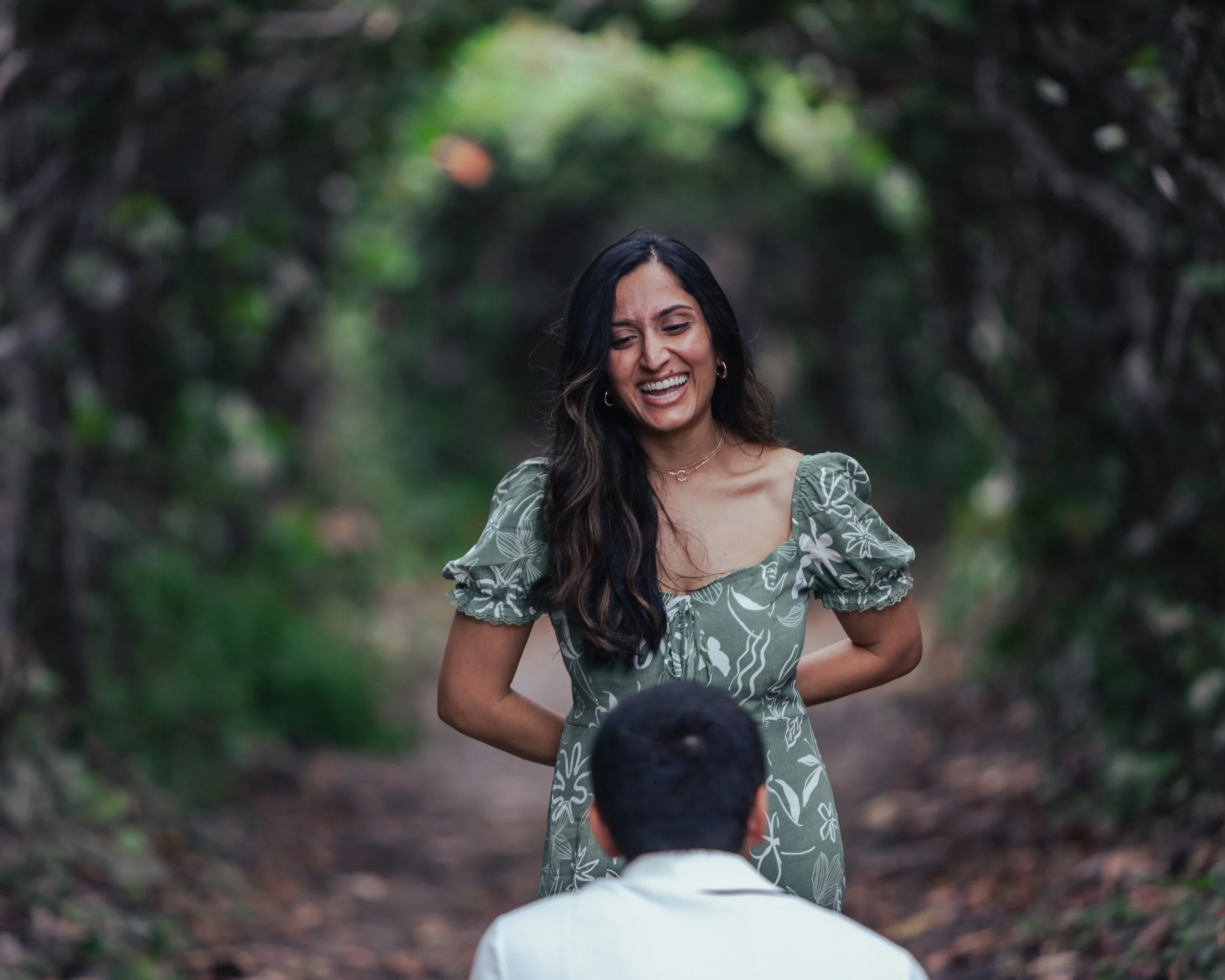 A woman with long dark hair smiling and standing outdoors on a forest path, her hands behind her back, wearing a green dress with white floral patterns. A man is kneeling in front of her with his back to the camera.