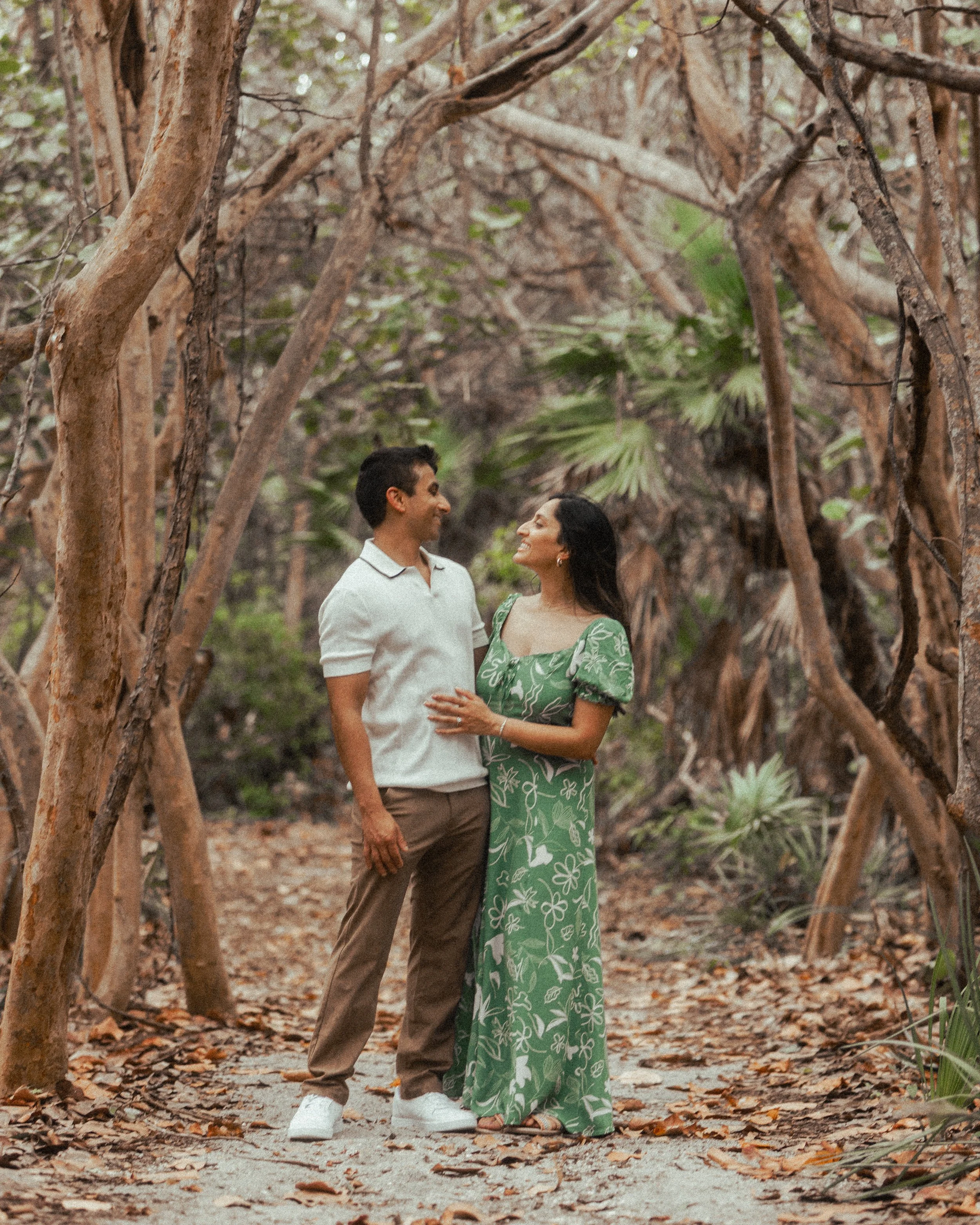 A couple standing together outdoors in a wooded area, smiling and looking at each other.
