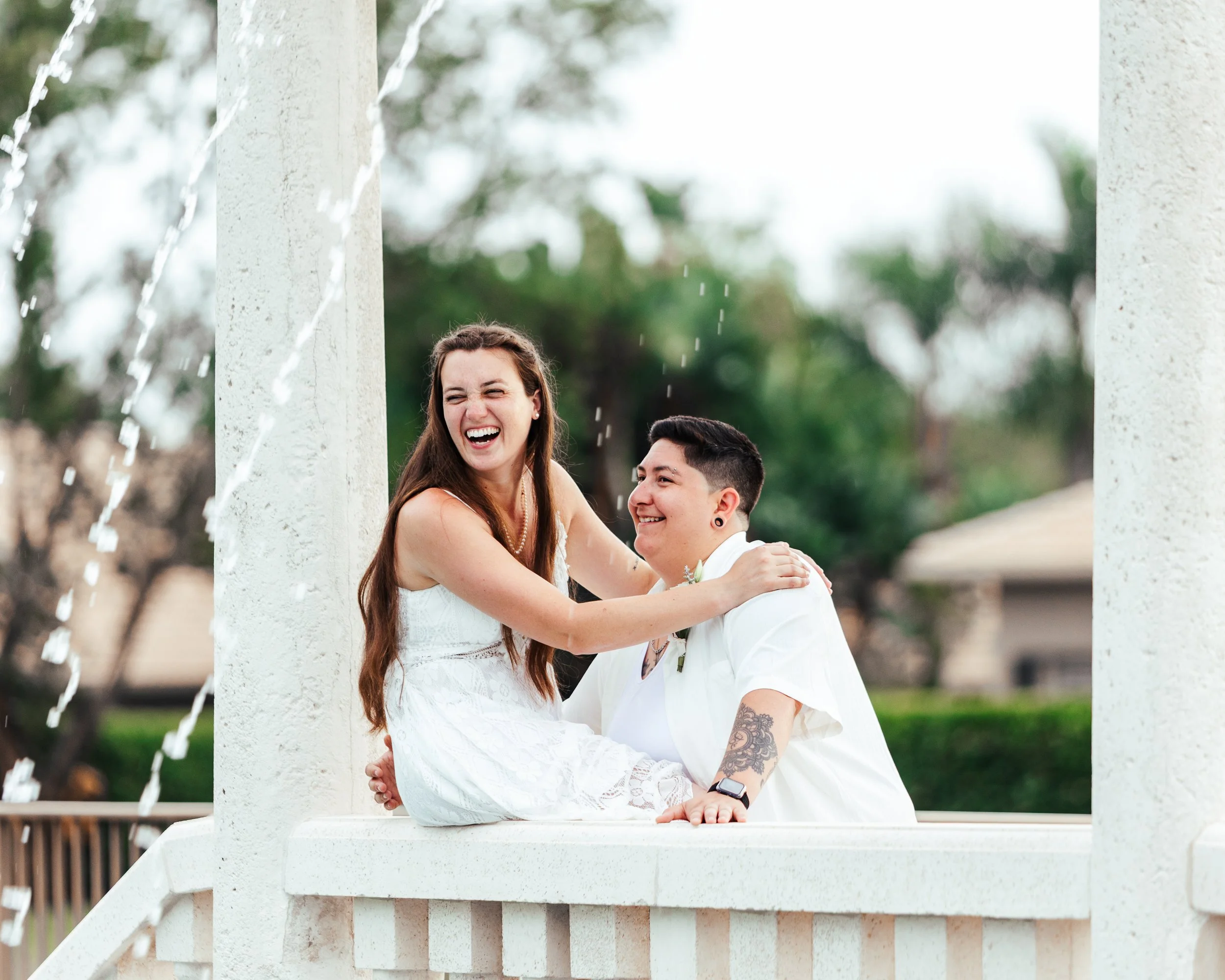 Two people sitting on a white fountain with water spraying, smiling and laughing outdoors.
