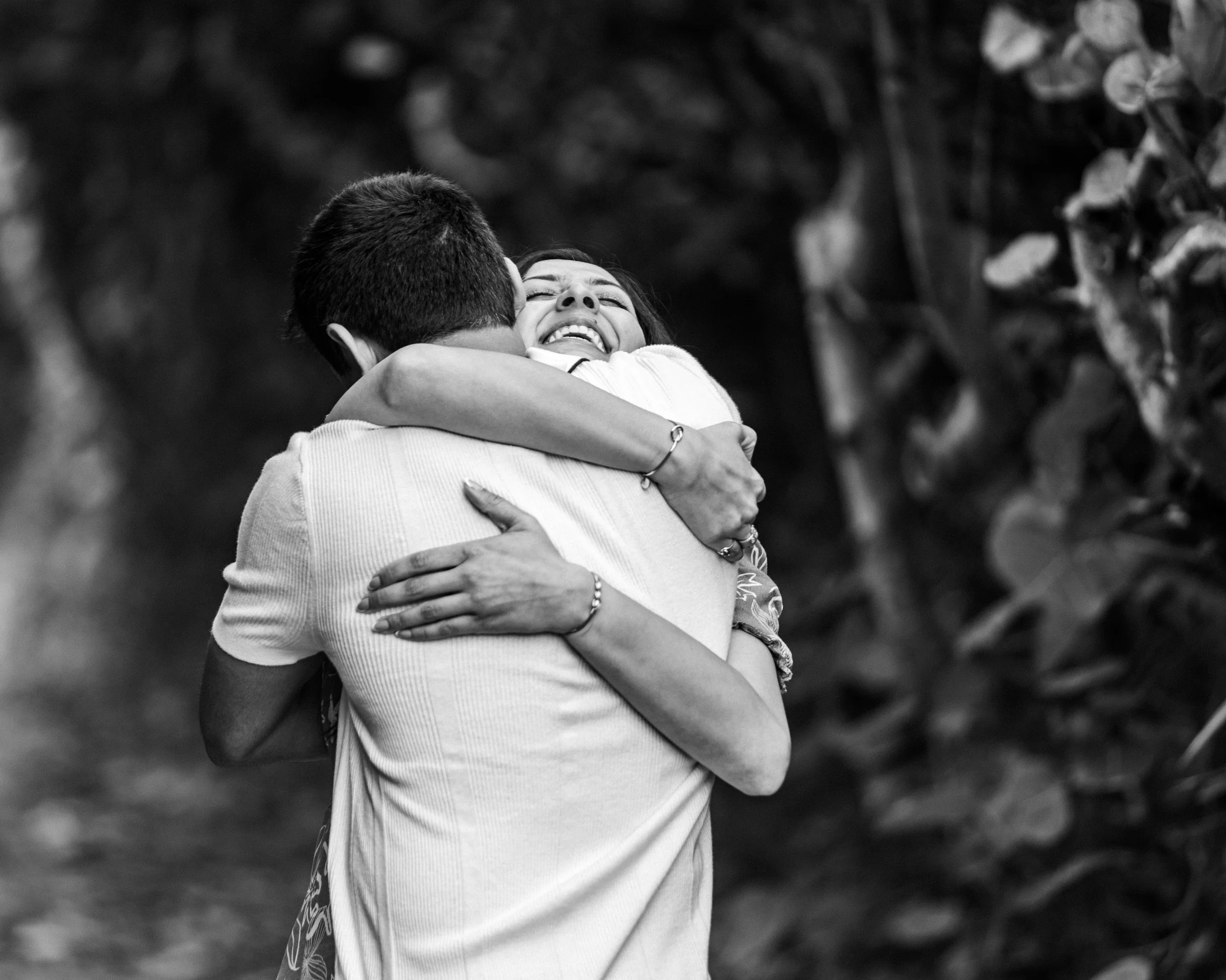 A black and white photo of a woman smiling happily as she hugs a man. They are outdoors with blurred foliage in the background.