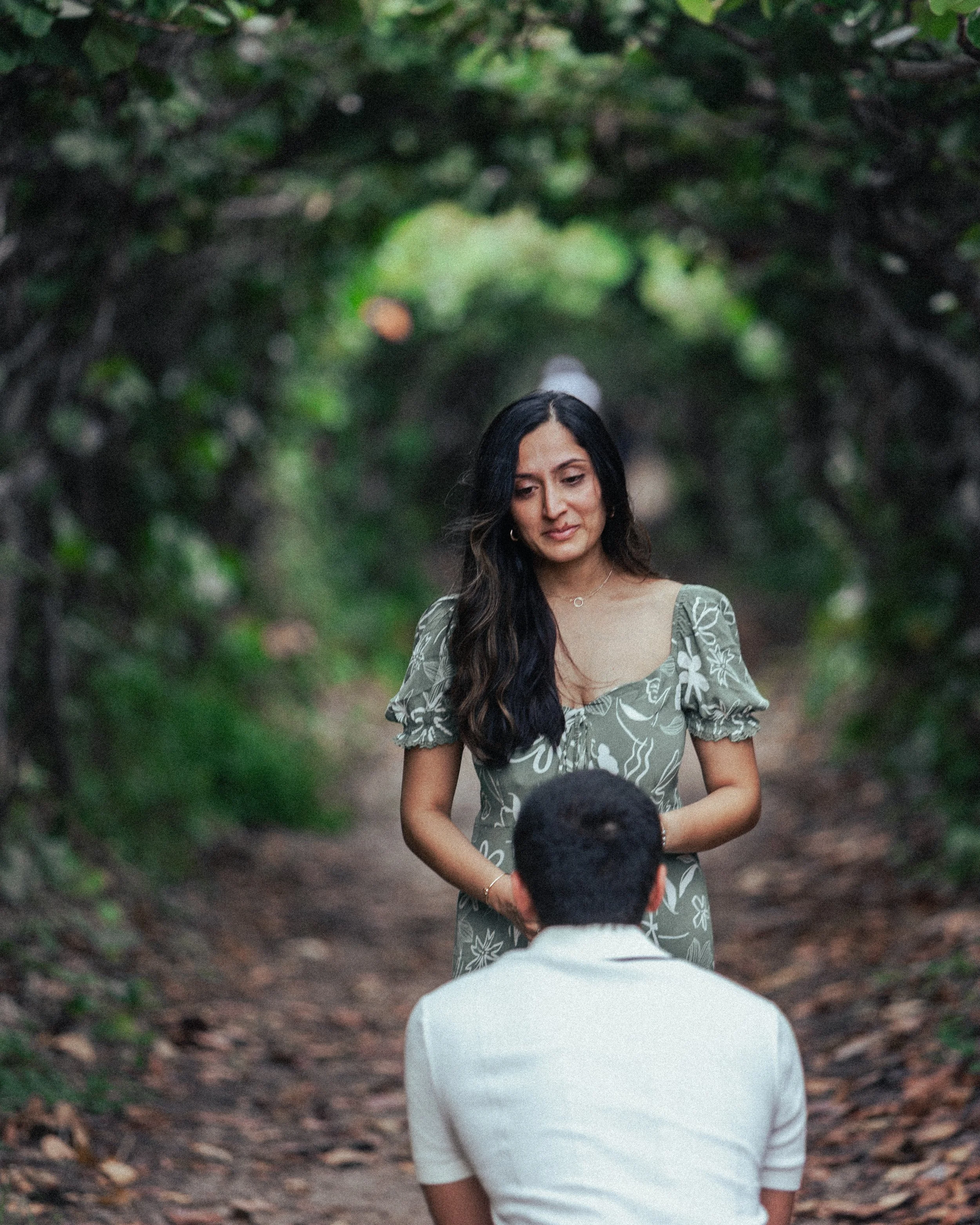 A woman proposing to a man on a forest trail, surrounded by lush greenery.