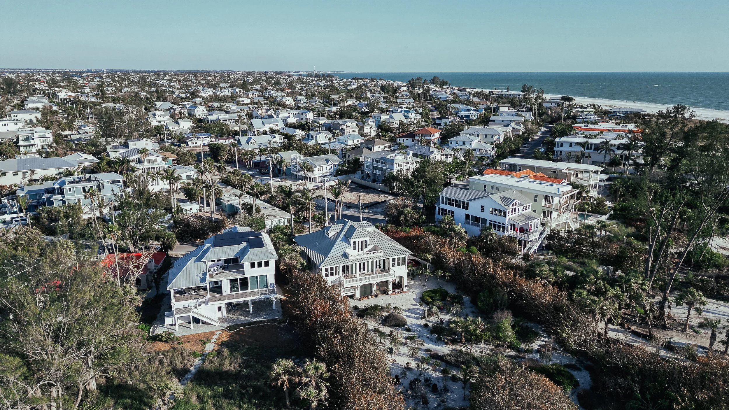 Aerial view of a coastal neighborhood with numerous houses, some with large porches and decks, situated among trees and greenery, with the ocean visible in the background.