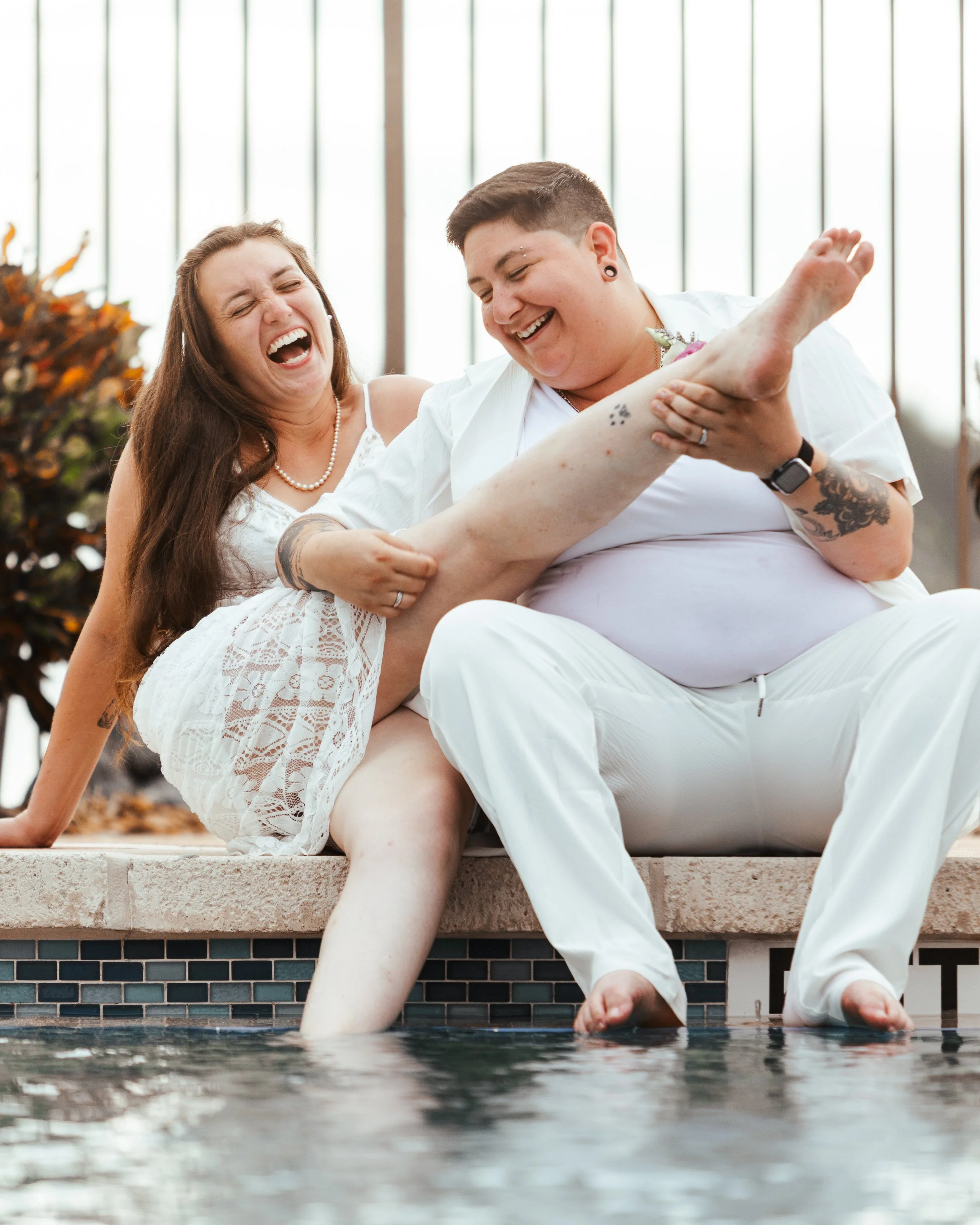 Two people sitting by a pool, smiling and laughing, holding one person's leg.