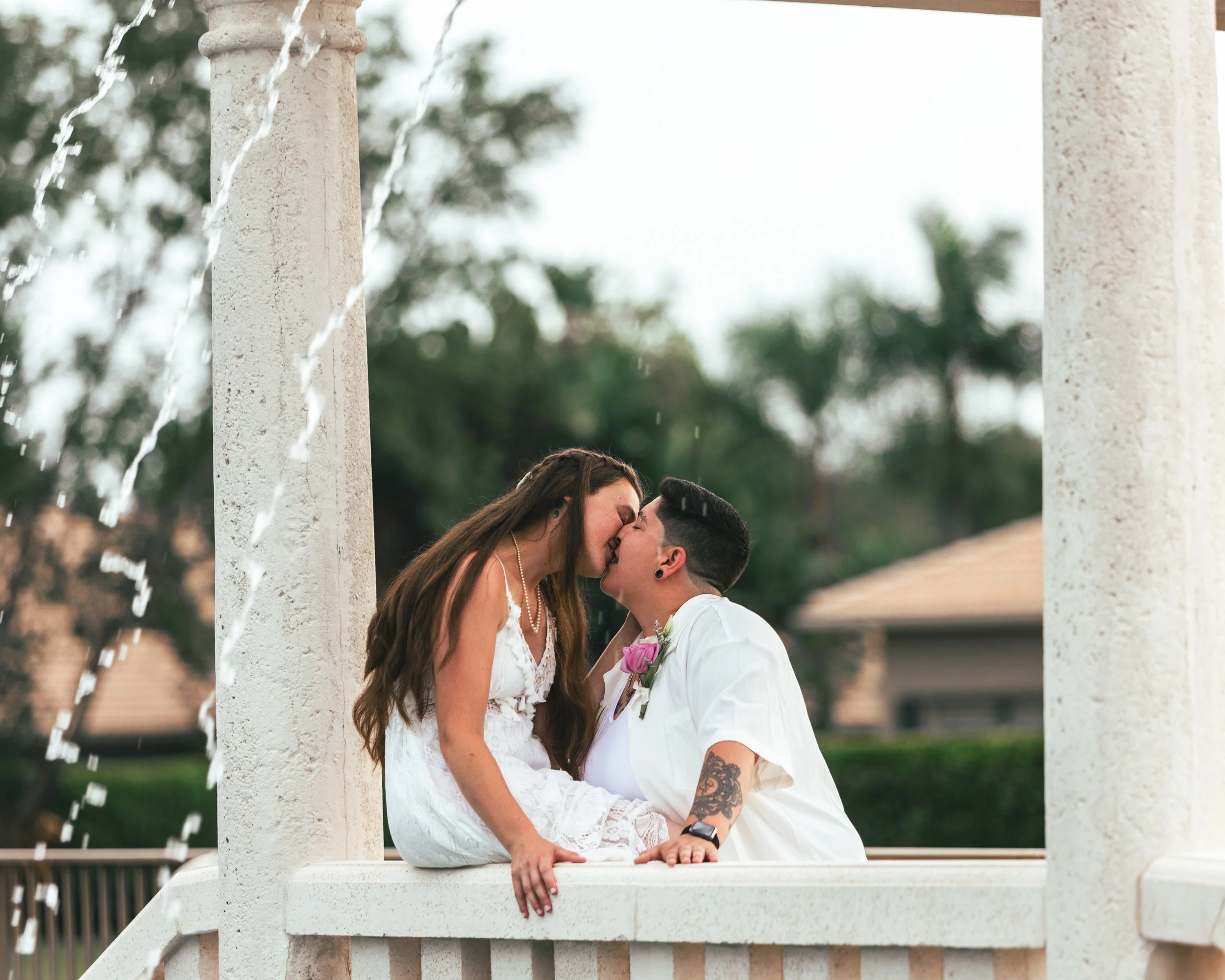 Couple kissing on a white bridge with water fountains and trees in the background.