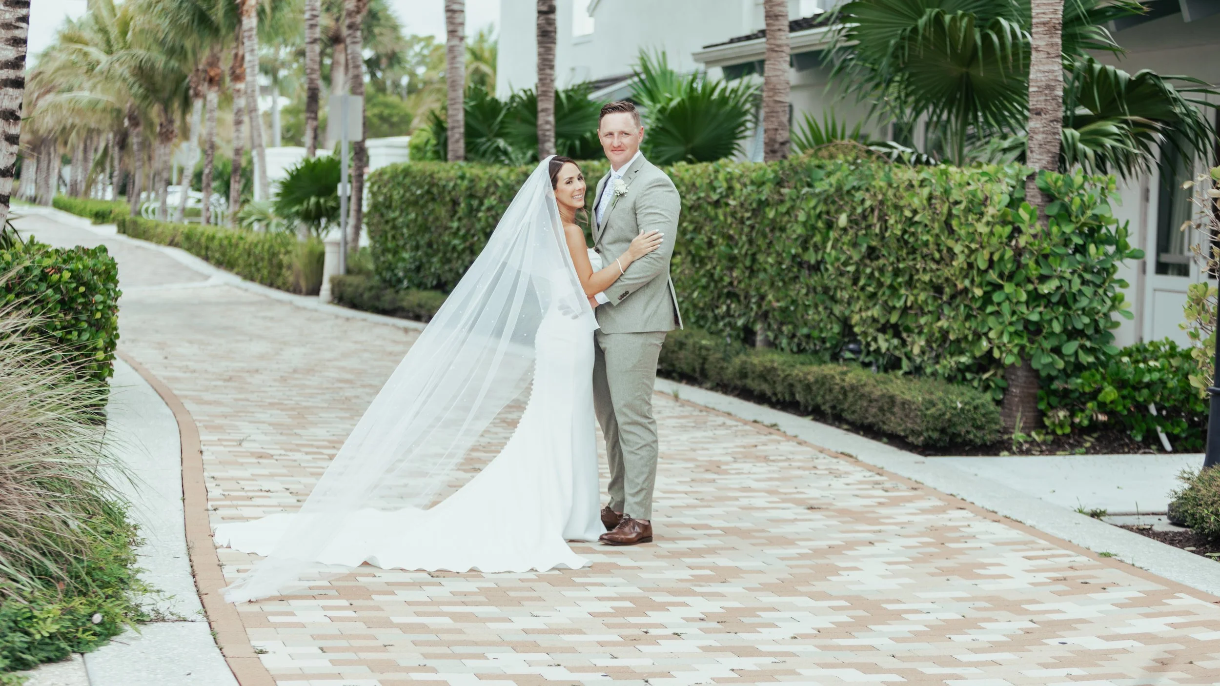 A bride and groom stand together on a brick pathway outdoors, surrounded by lush green bushes and palm trees during daytime.
