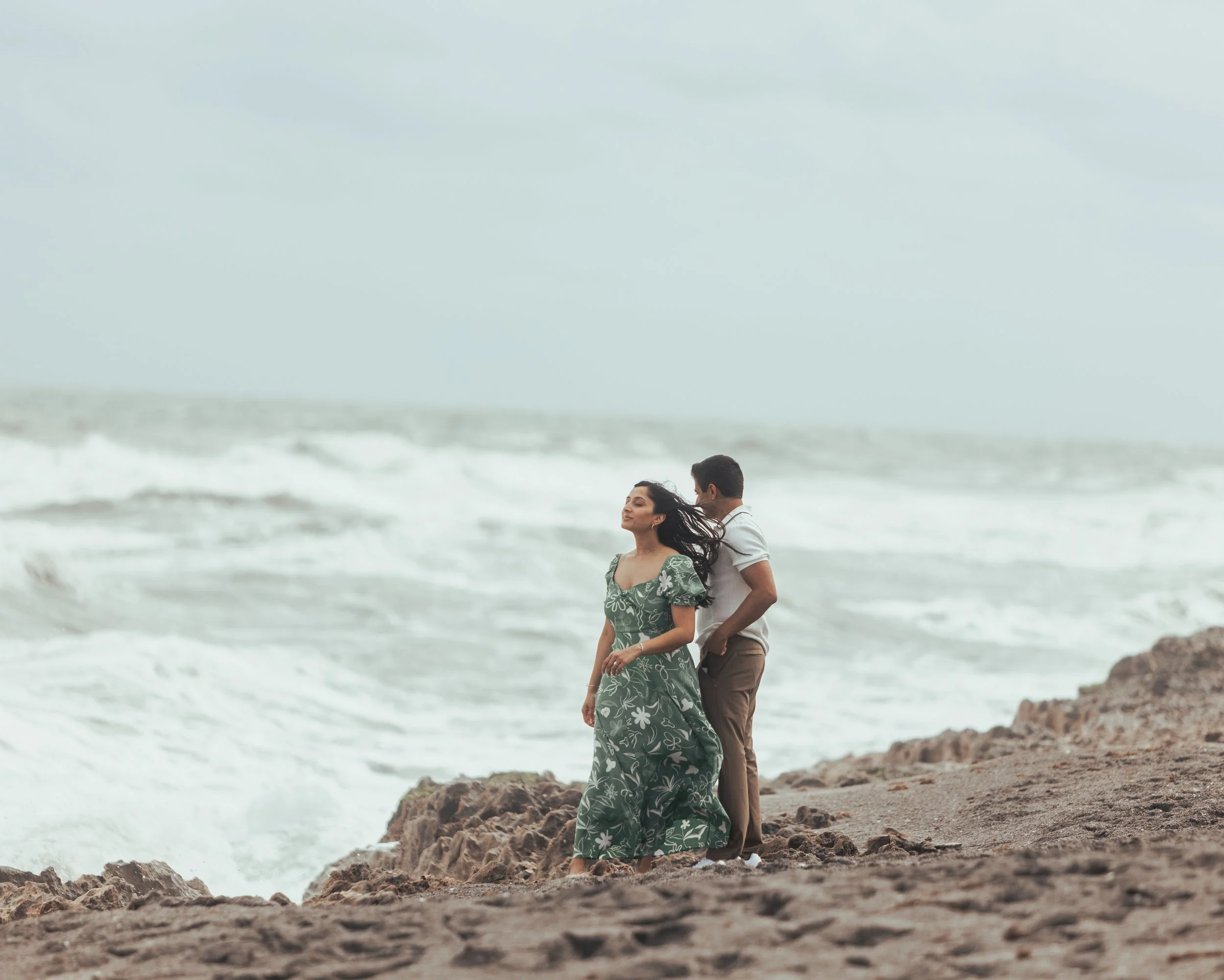 A couple standing on a rocky beach near the ocean, with waves in the background, one woman in a green floral dress and a man in a white shirt and brown pants.