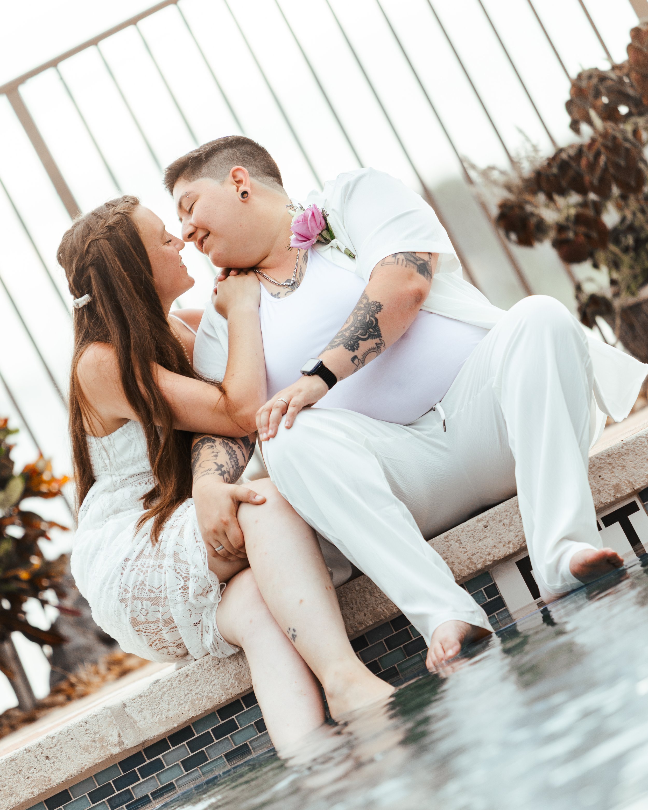 A couple sitting by a pool, embracing and smiling at each other, with water and plants in the background.