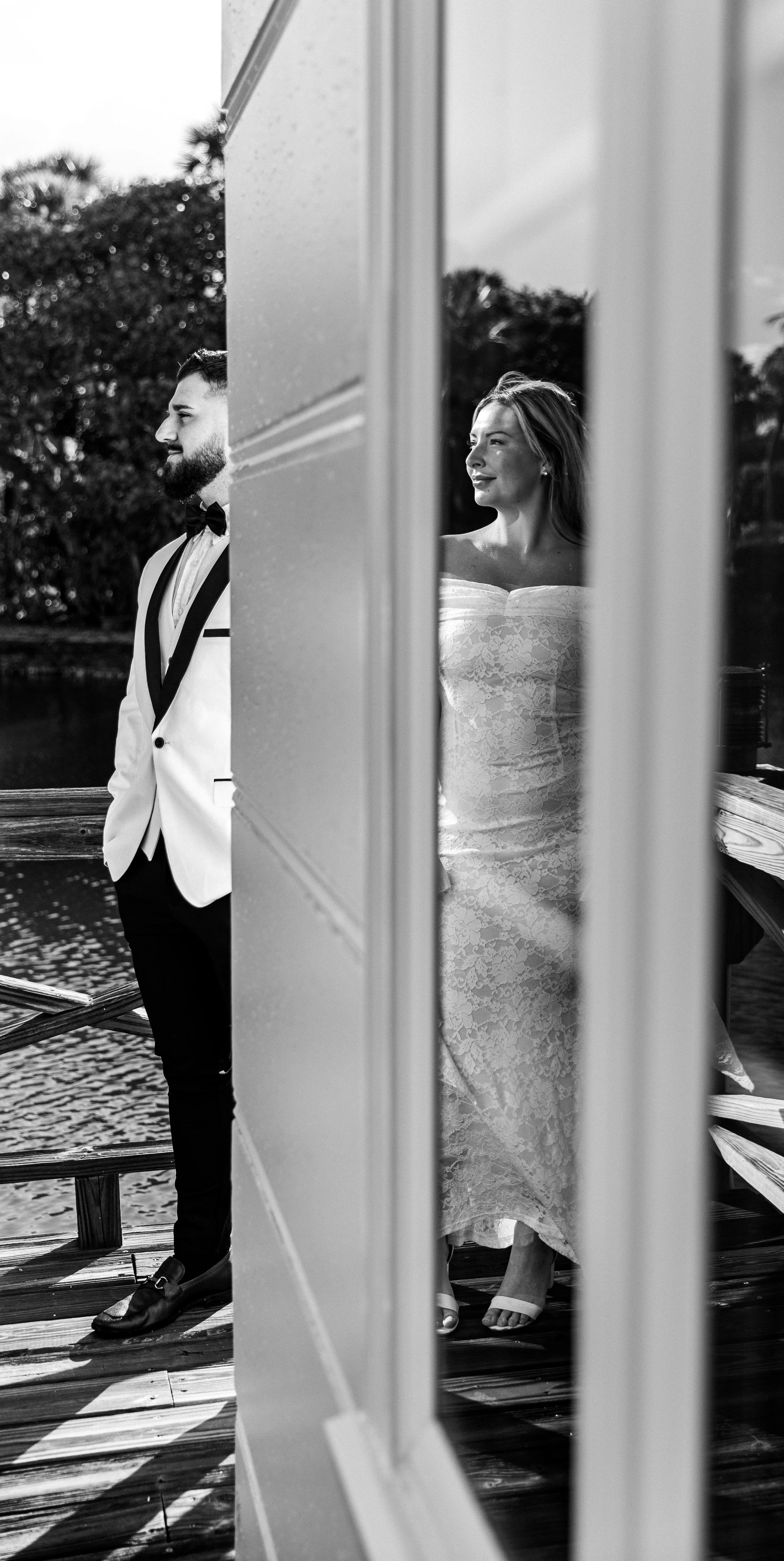 A black and white photo of a bride and groom standing outside near a body of water, partially visible behind a wall or partition, with trees in the background. The groom, on the left, wears a white tuxedo jacket with a black bow tie and black pants, looking to the side. The bride, on the right, wears a strapless lace wedding dress and looks at the groom. The photo is taken from outside, through a window or door, framing the couple.